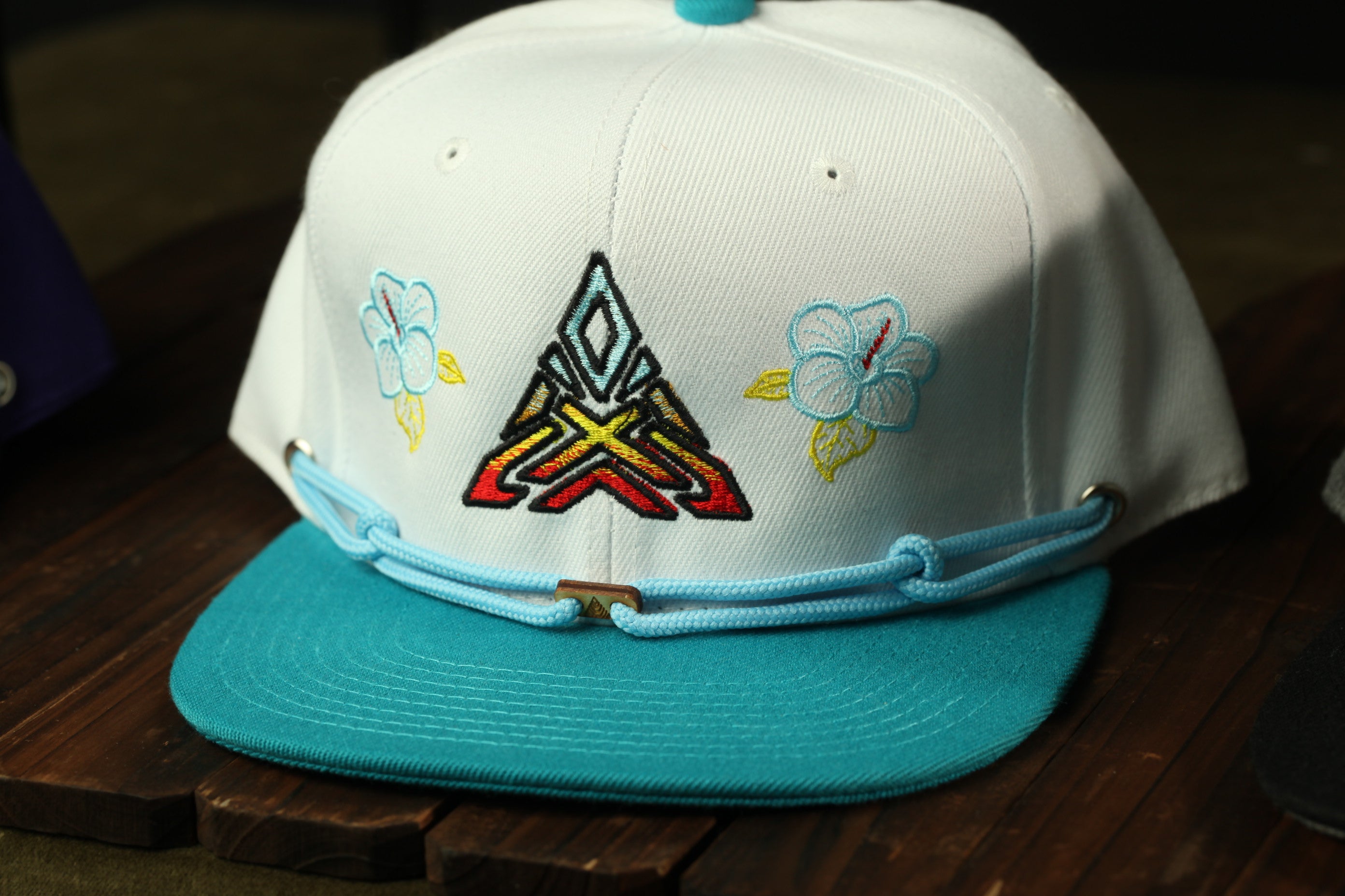 A white baseball cap with colorful floral embroidery and a blue brim, displayed on a wooden surface.