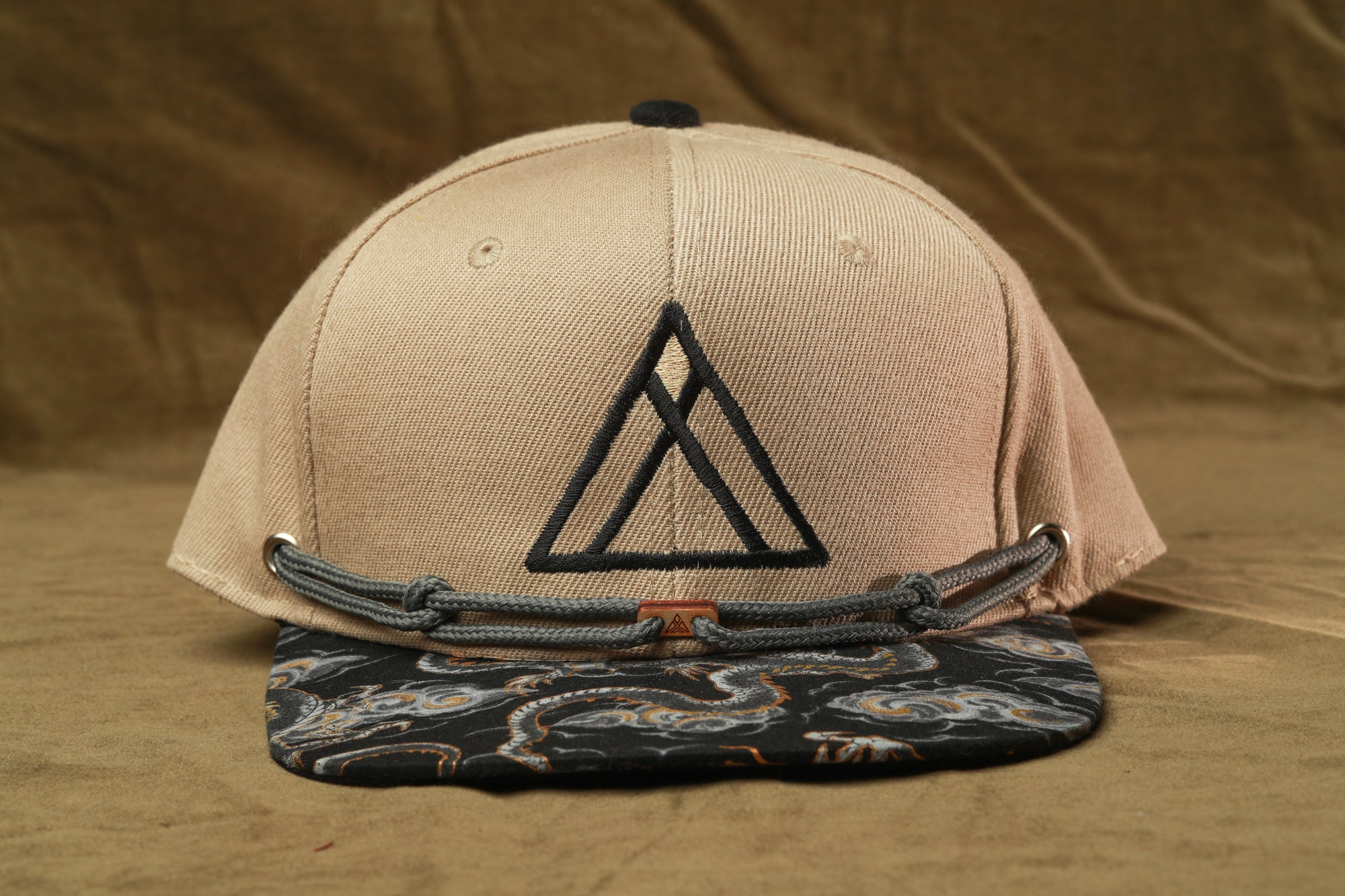 A beige baseball cap with a black triangle logo on the front, placed on a brown fabric background.