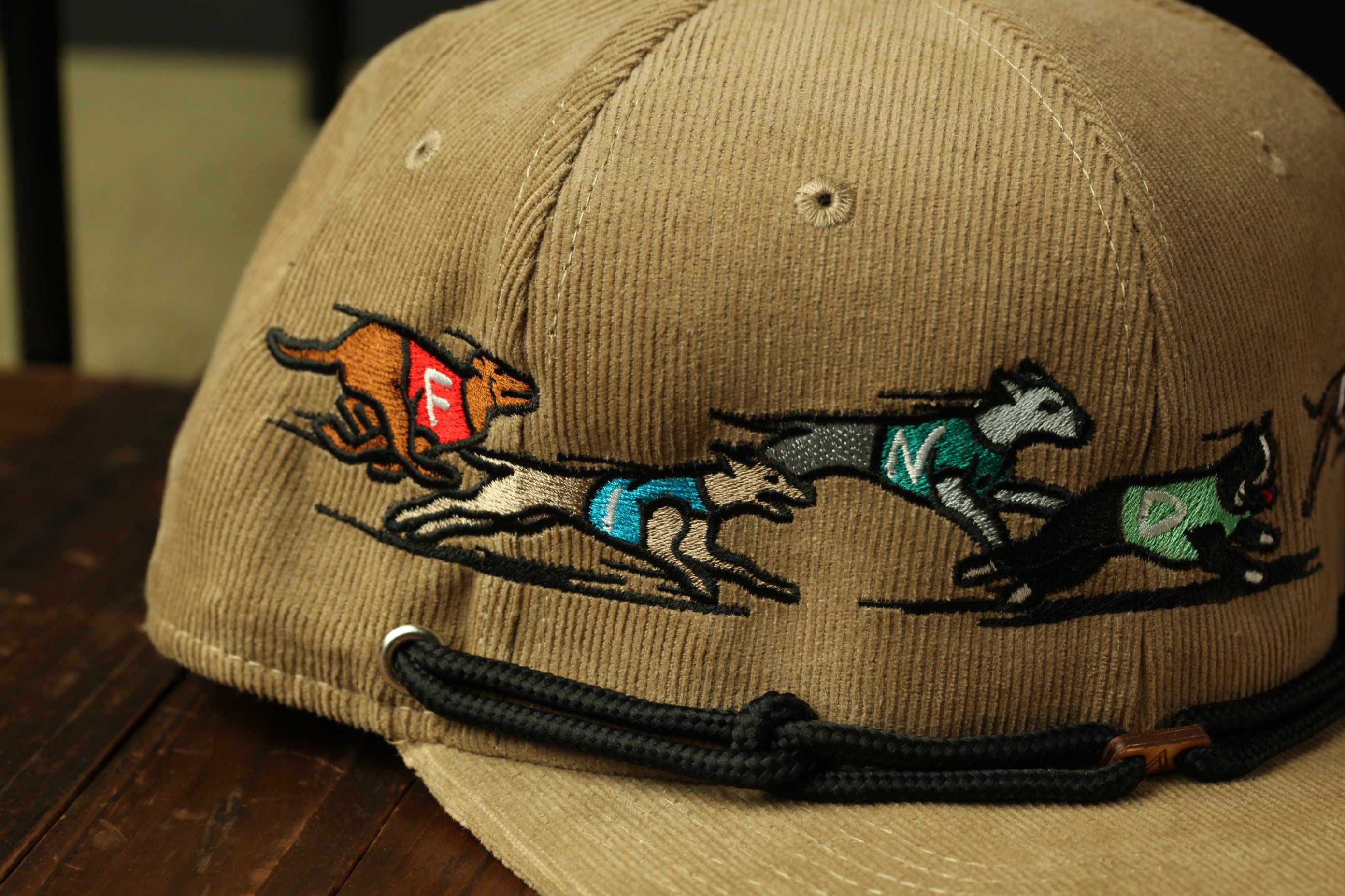 A beige baseball cap with a detailed embroidered design depicting three dogs in motion, with one dog wearing a red vest.