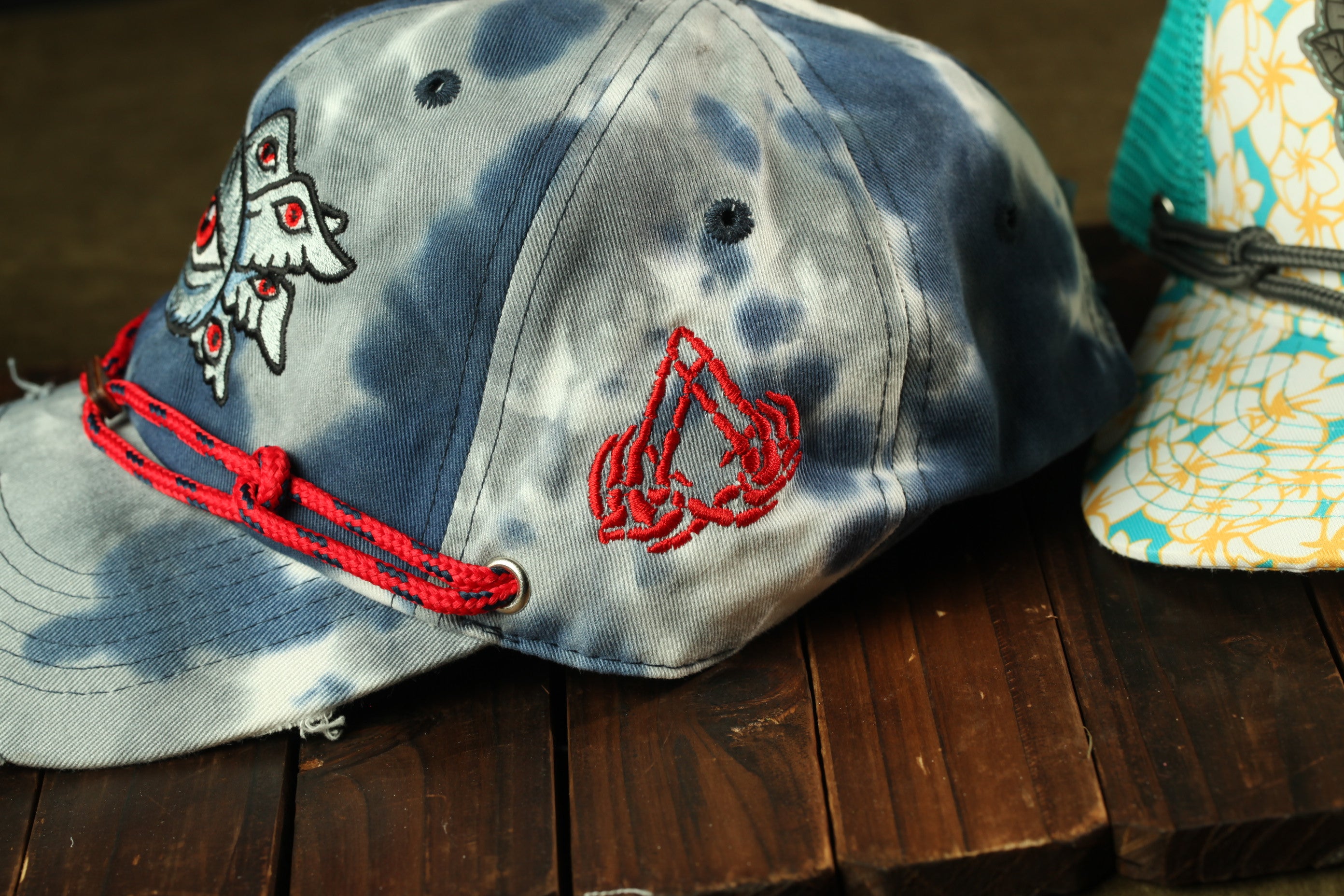 A blue and gray baseball cap with red embroidered designs, including a butterfly and a skull, is shown on a wooden surface.
