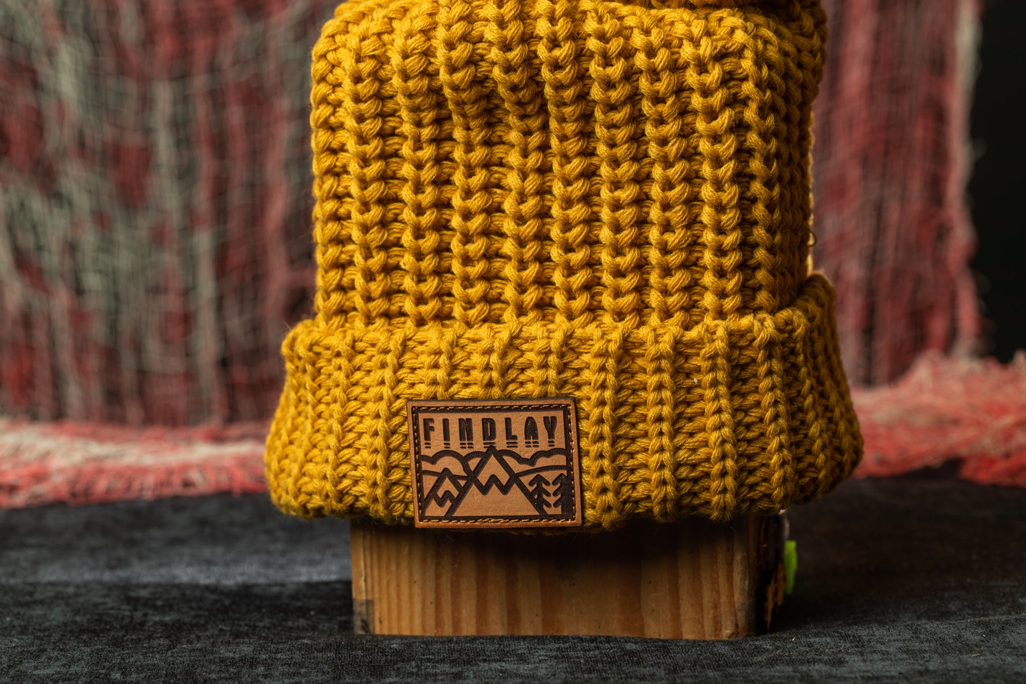 A yellow knit beanie with a wooden block in the center, featuring a leather patch with a mountain design.
