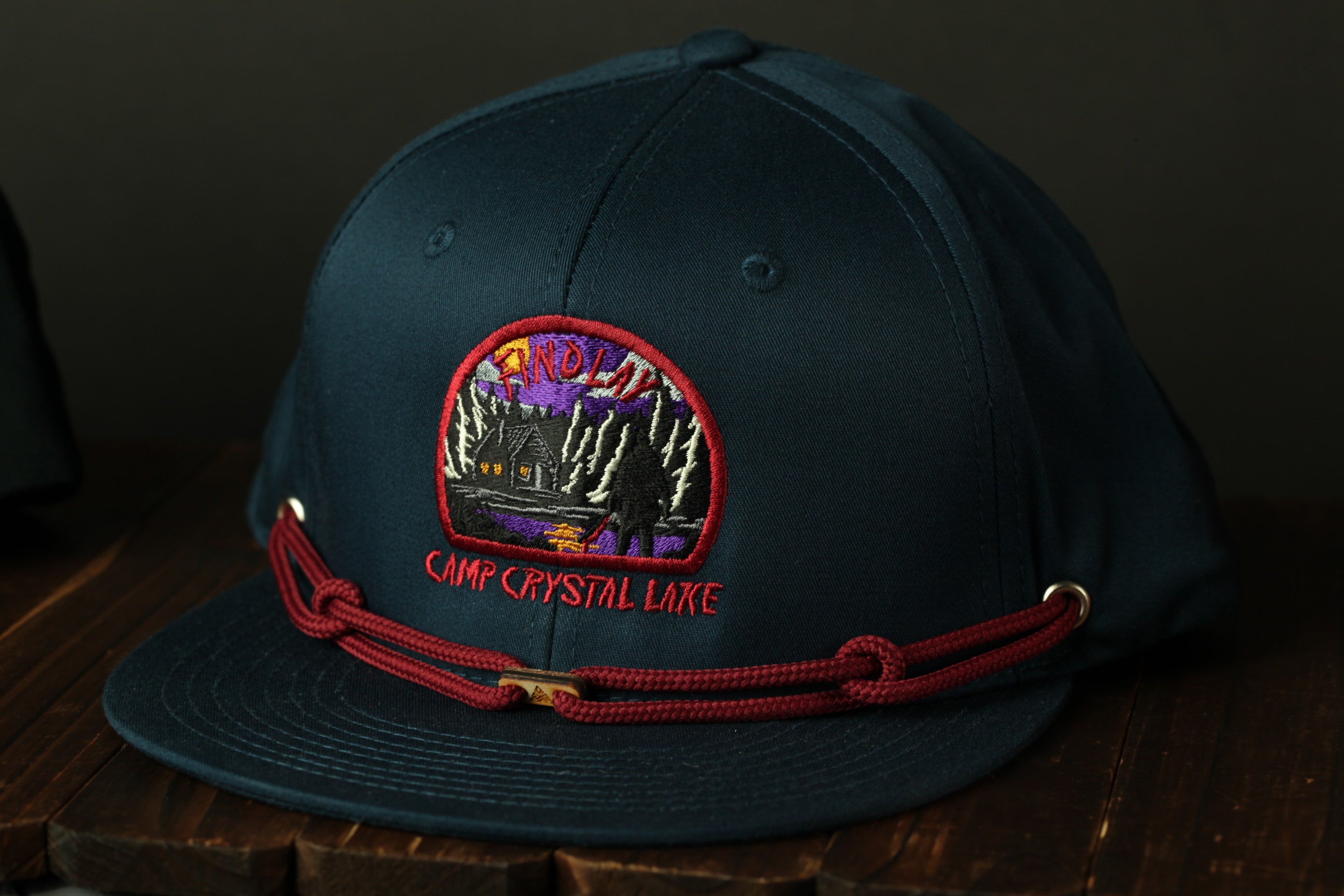 A dark blue baseball cap with a red string and a circular embroidered patch on the front, featuring a scene from Camp Crystal Lake.