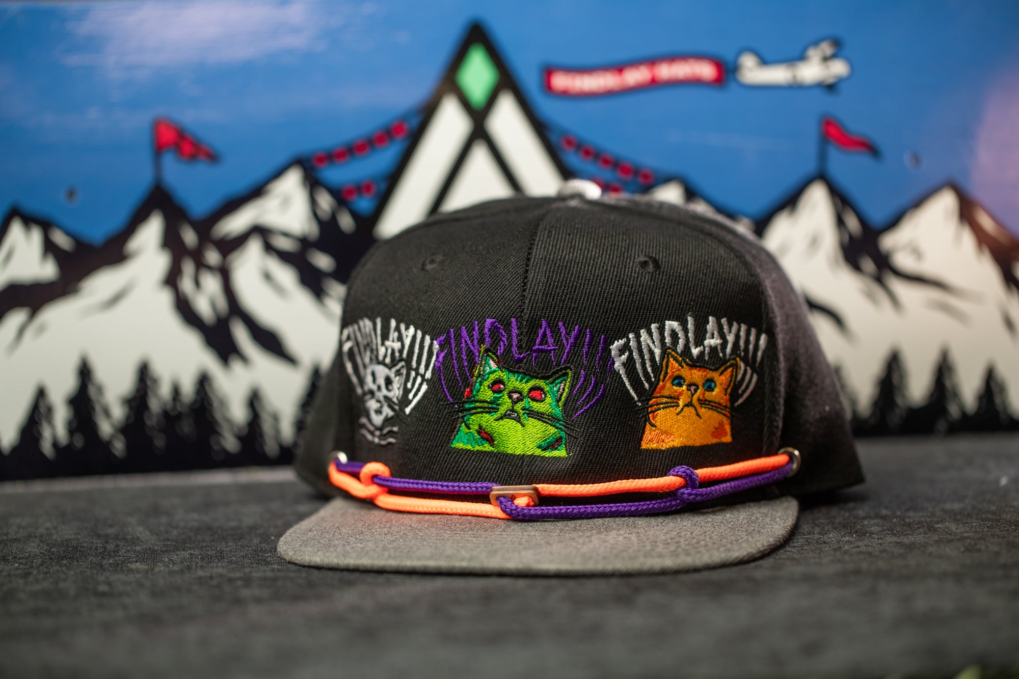 A black baseball cap with colorful cat designs and text on it, placed on a gray surface against a background of mountains and flags.