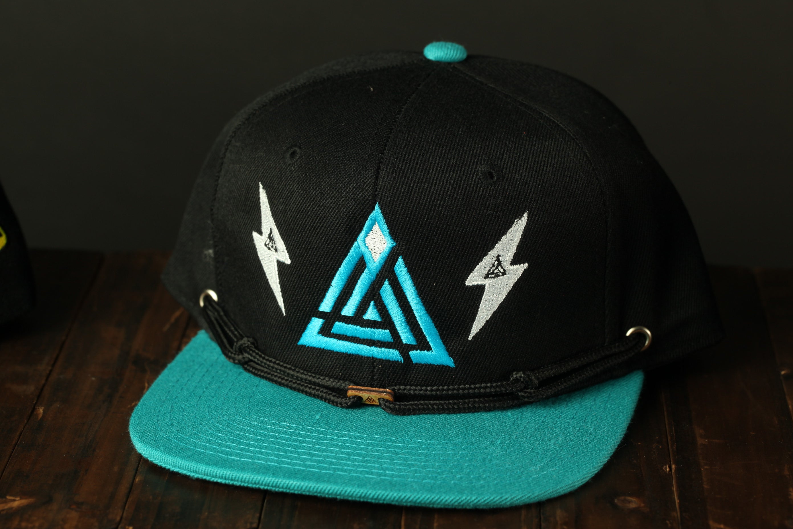 A black baseball cap with a teal brim and a geometric design on the front, placed on a wooden surface.