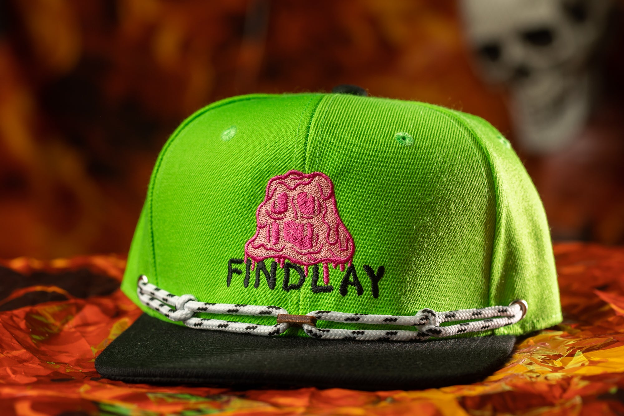 A bright green baseball cap with a pink graphic and the word "FINDLAY" printed on it, placed on a red and orange patterned surface.