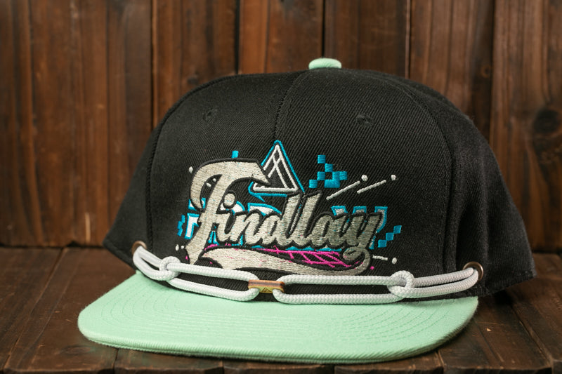 A black baseball cap with a teal brim and a graphic design on the front, placed on a wooden surface.