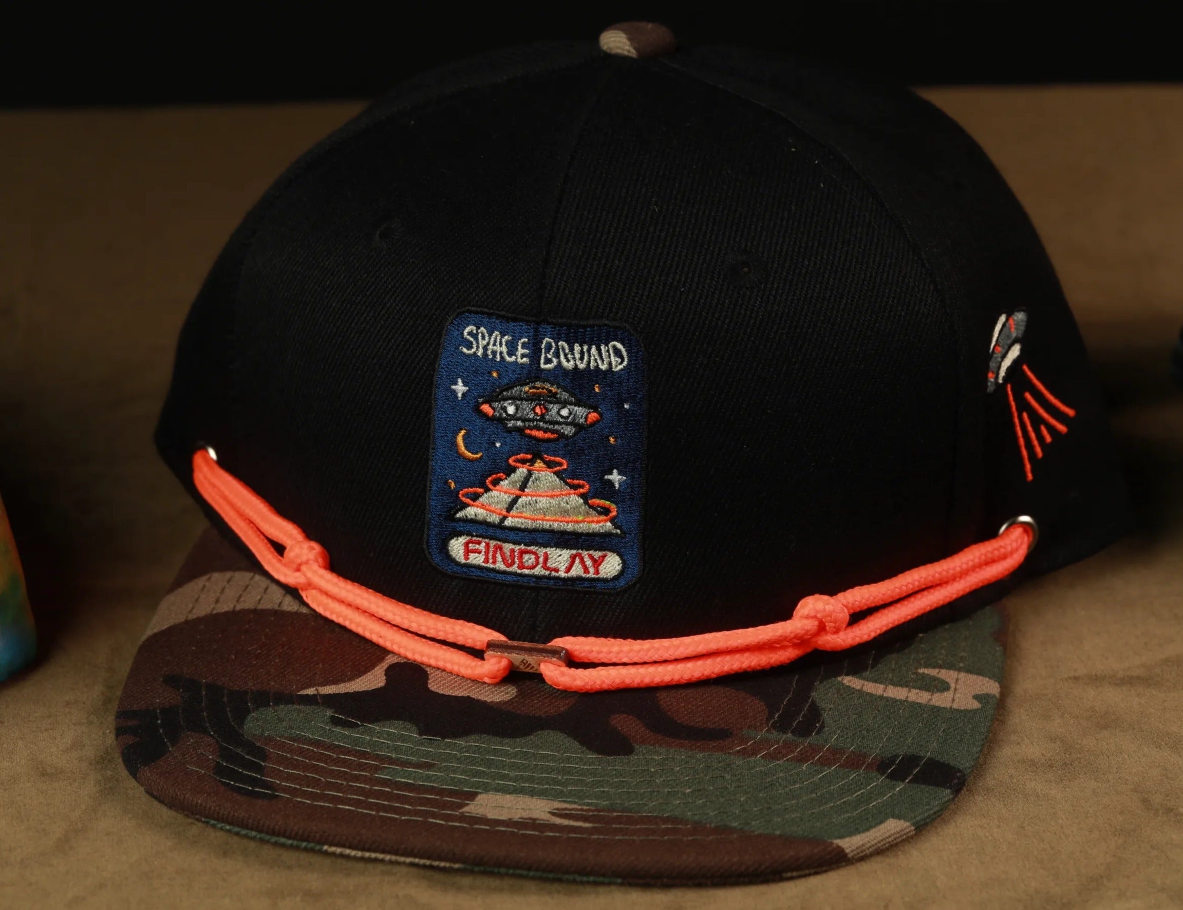 A black baseball cap with a camouflage pattern and orange strings, featuring a patch with the text "Space Bound" and an illustration of an alien spacecraft.
