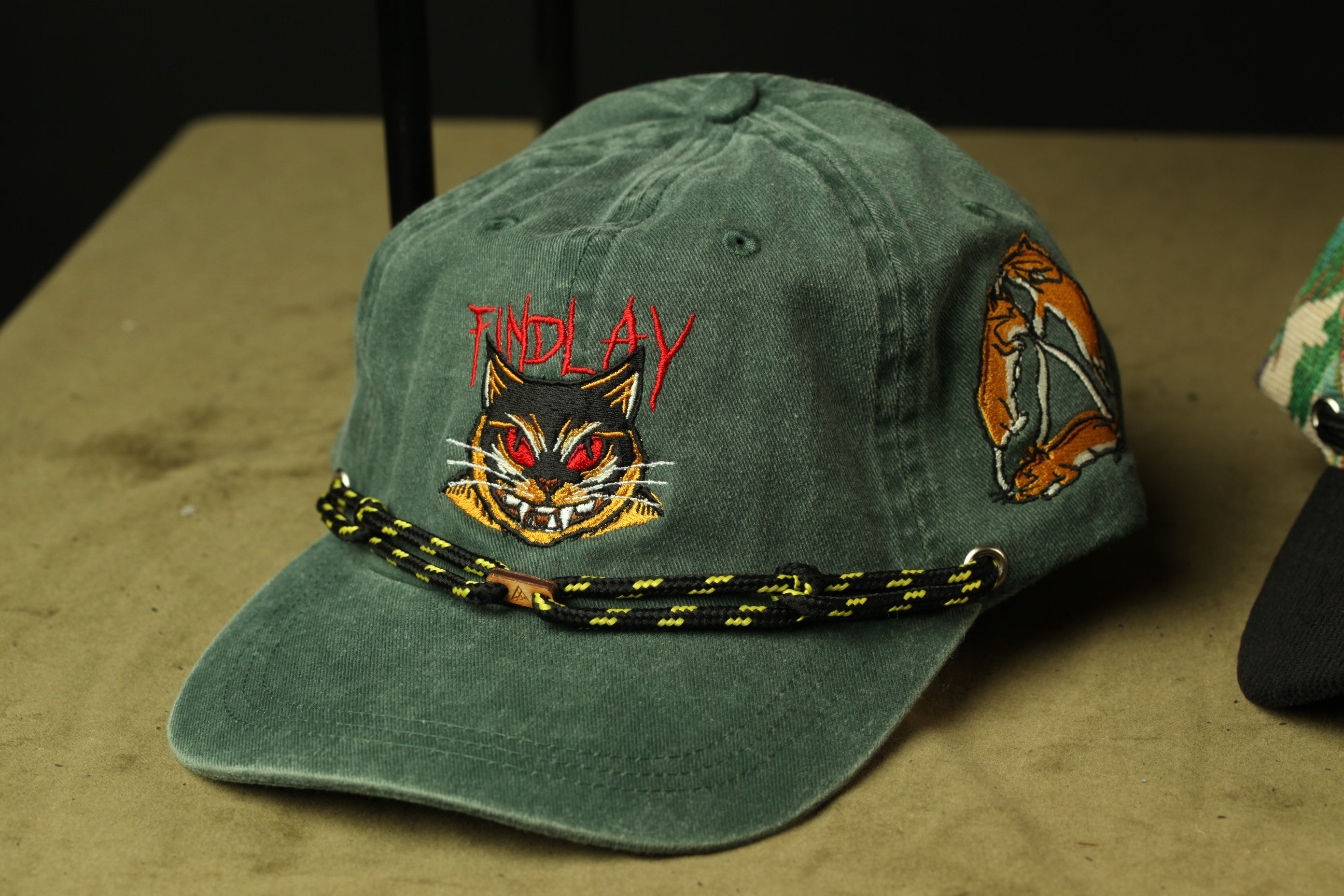 A green baseball cap with a cat face design and the word "FINDLAY" printed on it.