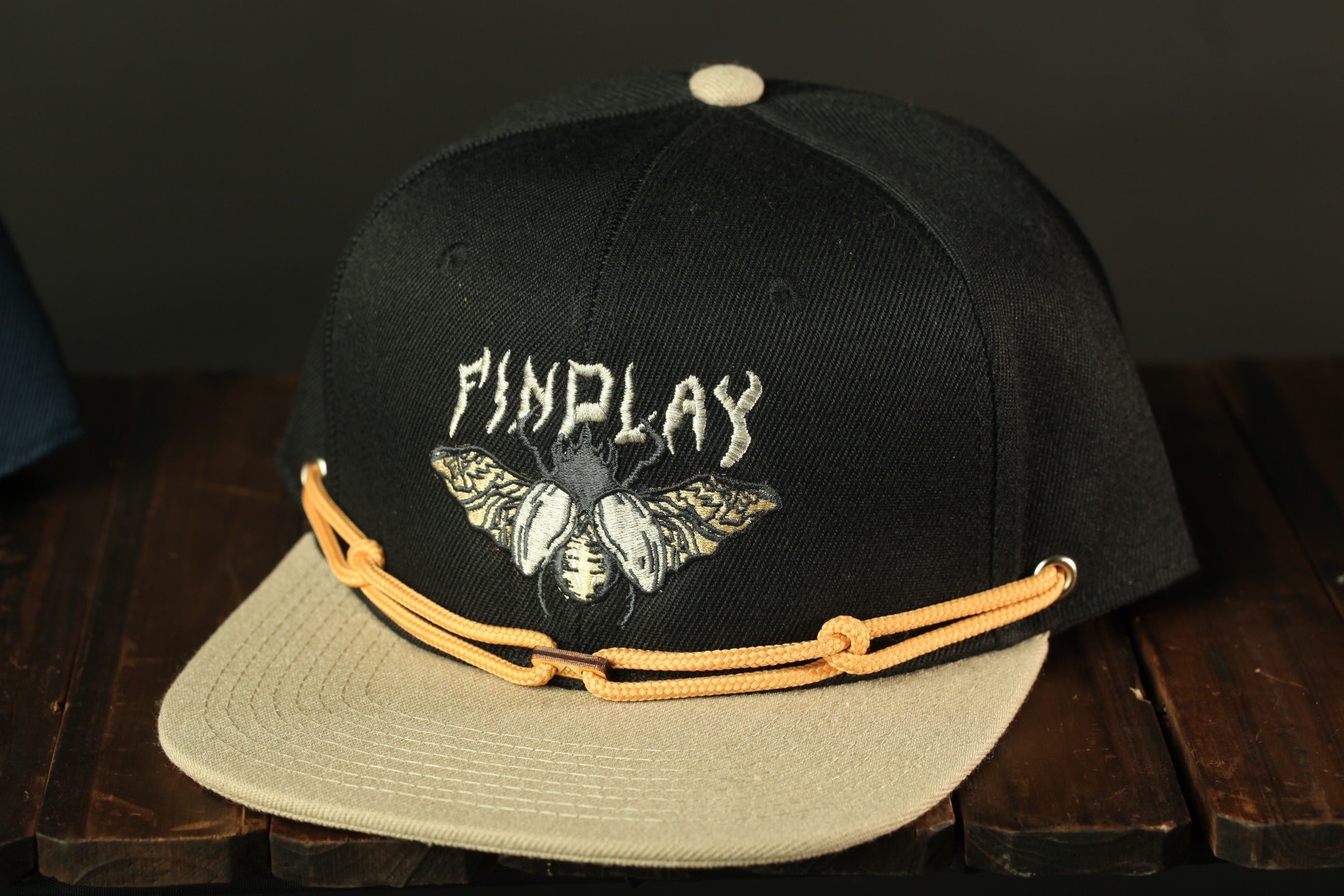 A black baseball cap with a beige brim and a white logo on the front, sitting on a wooden surface.