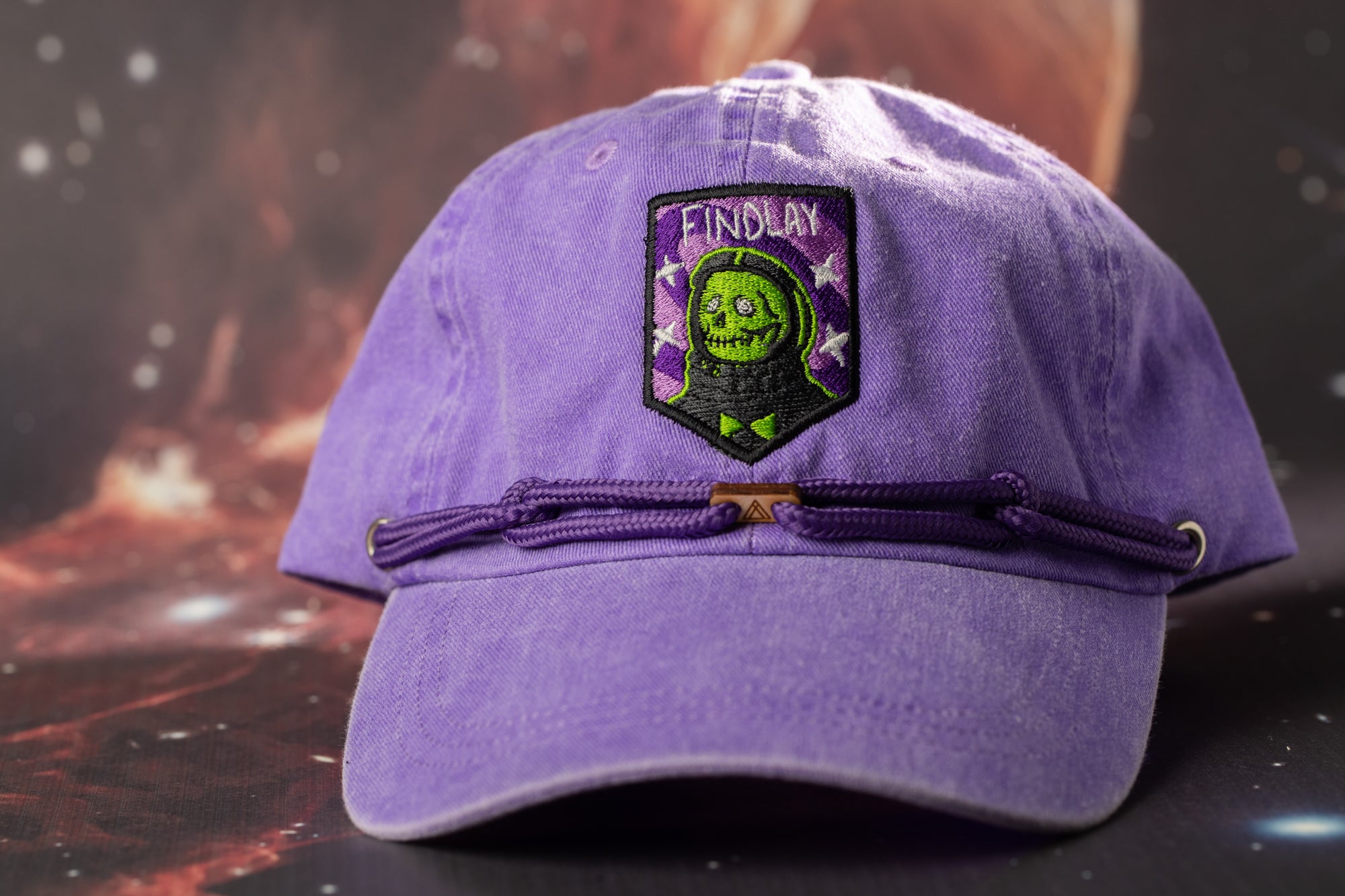A purple baseball cap with a green skull and crossbones design on the front, featuring a patch with the text "FINDLAY" on it.