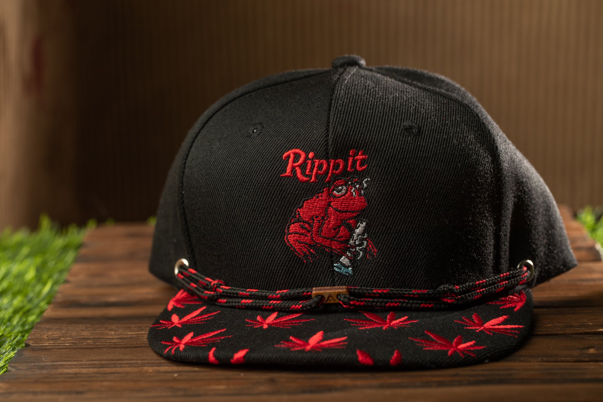 A black baseball cap with a red and black pattern and a red frog embroidered on the front, sitting on a wooden surface.
