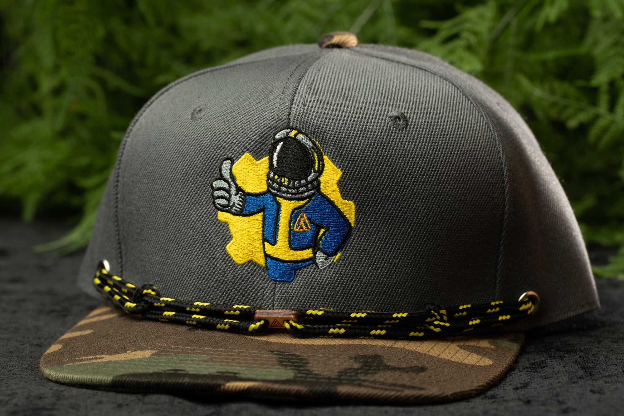 A gray baseball cap with a yellow and blue embroidered design depicting a person wearing a spacesuit, giving the impression of a space suit or astronaut.
