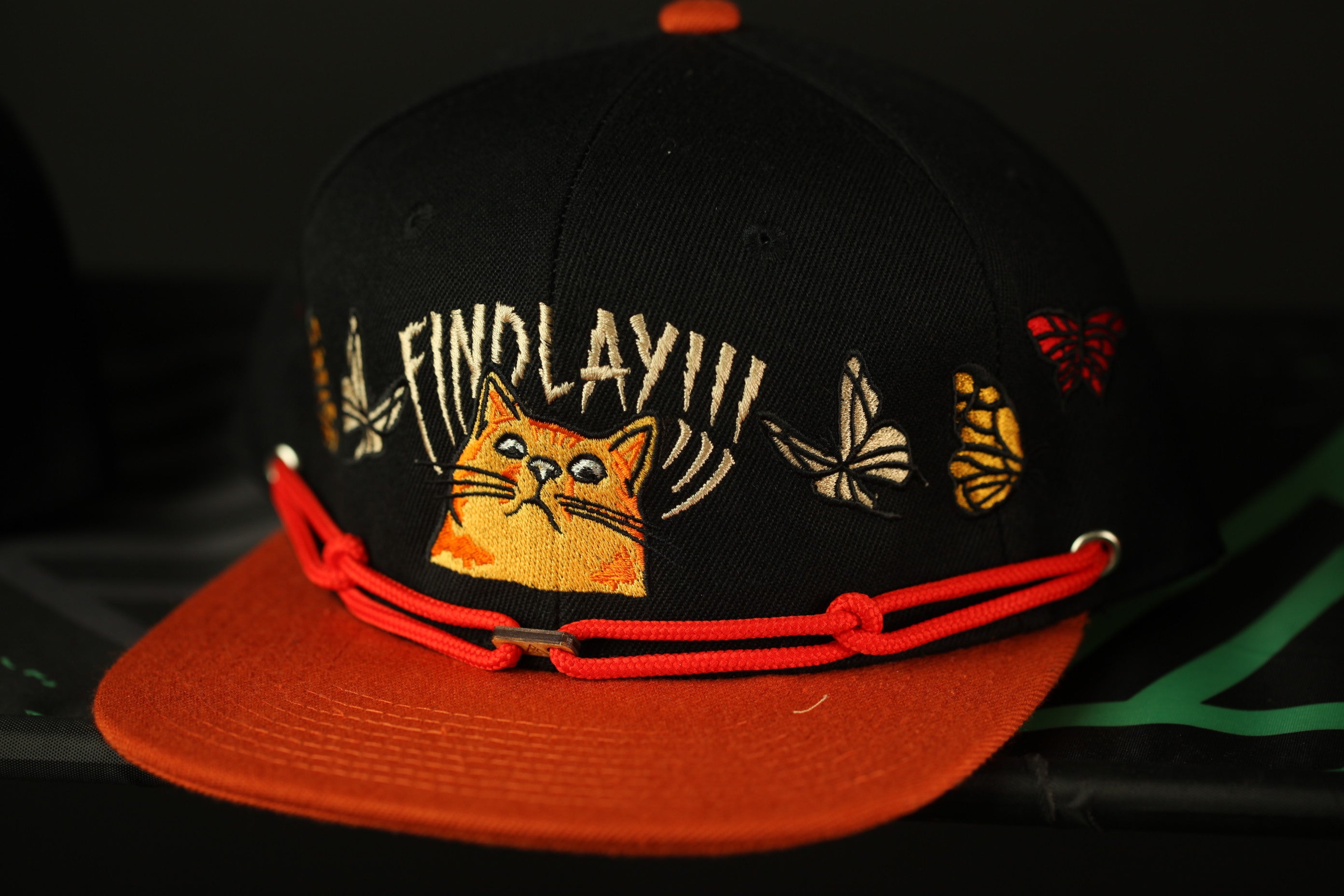 A black baseball cap with a red band and an embroidered cat design on it.