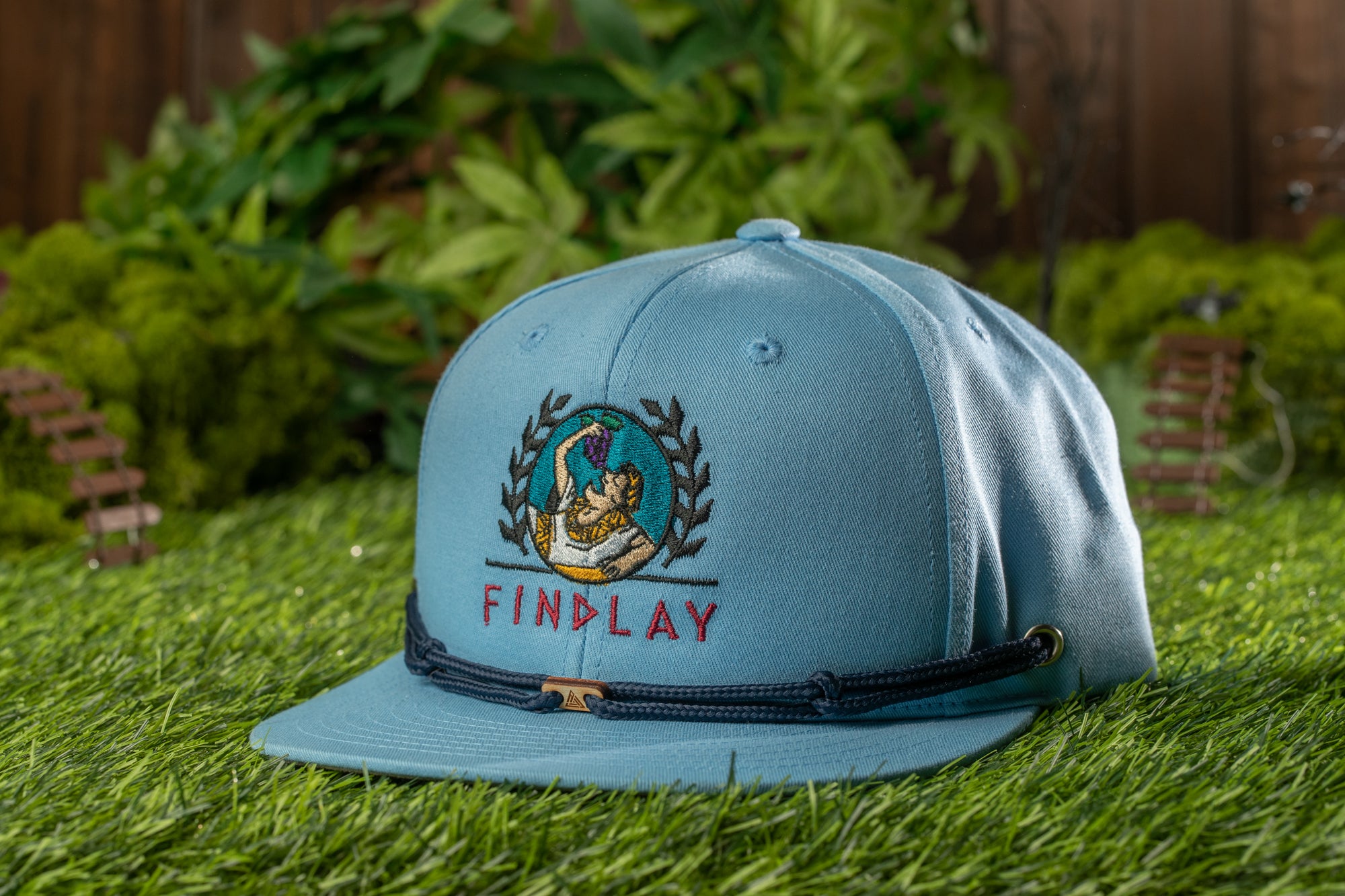 A light blue baseball cap with a logo and text on it, placed on a grassy surface with some small wooden structures in the background.