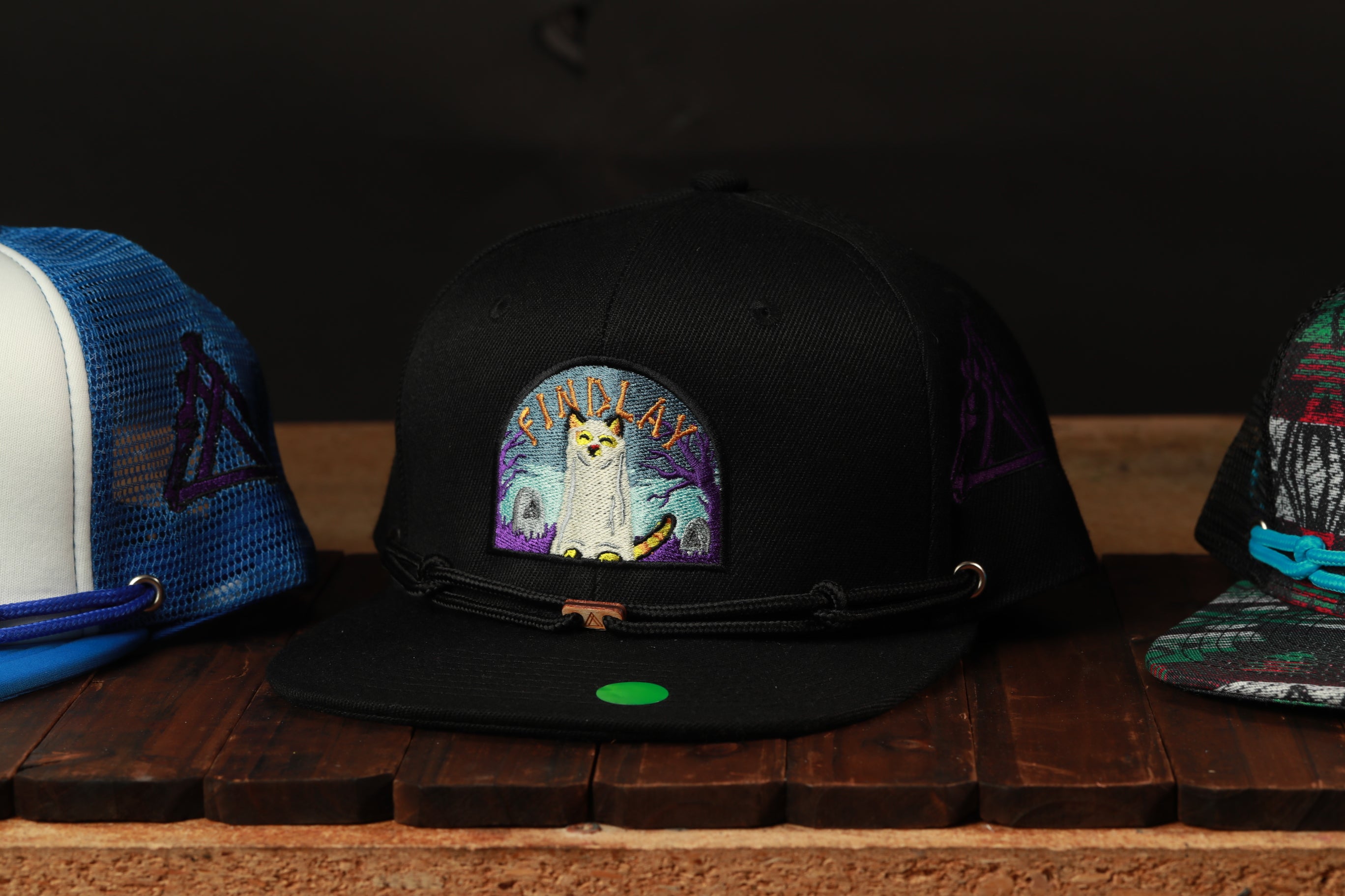A collection of colorful baseball caps is displayed on a wooden surface, with one black cap featuring a cat embroidered on it.