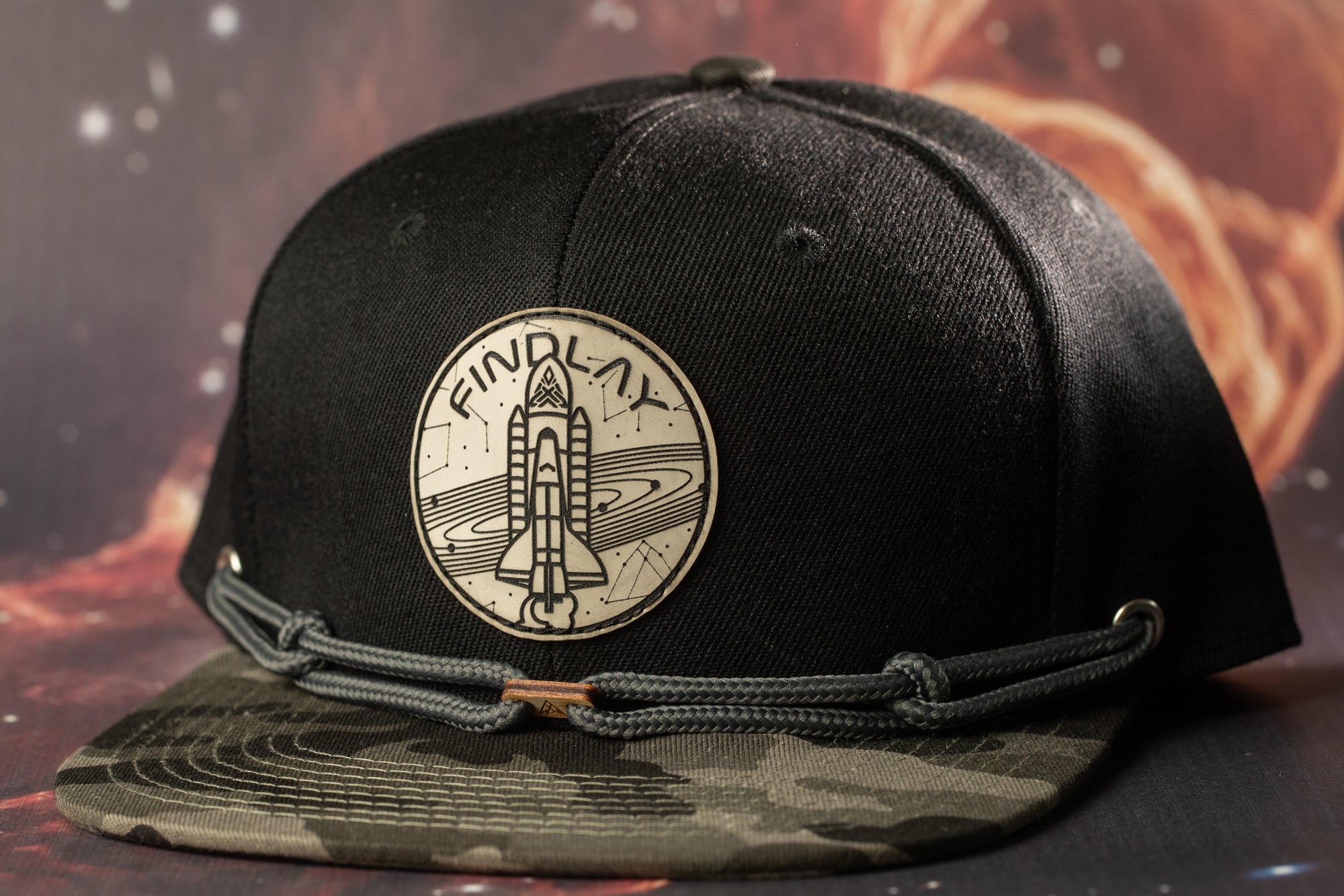 A black baseball cap with a space shuttle motif and the word "FINDLAY" on it, set against a background of a space-themed design.