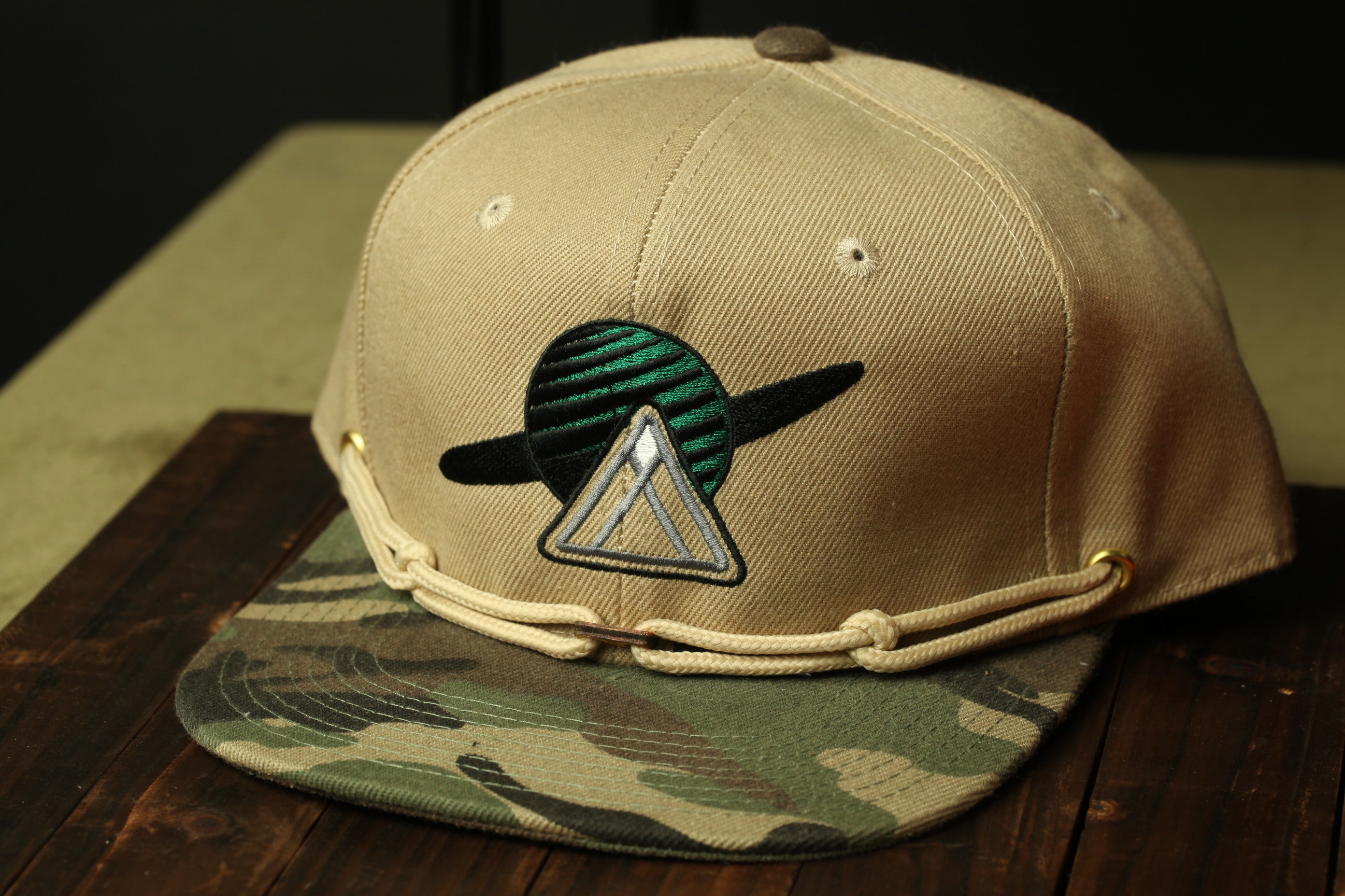 A beige baseball cap with a camouflage pattern and a triangular patch on the front, placed on a wooden surface.