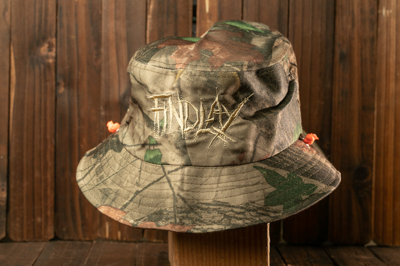 A camouflage bucket hat with the word "FIND" embroidered on it, displayed on a wooden surface.