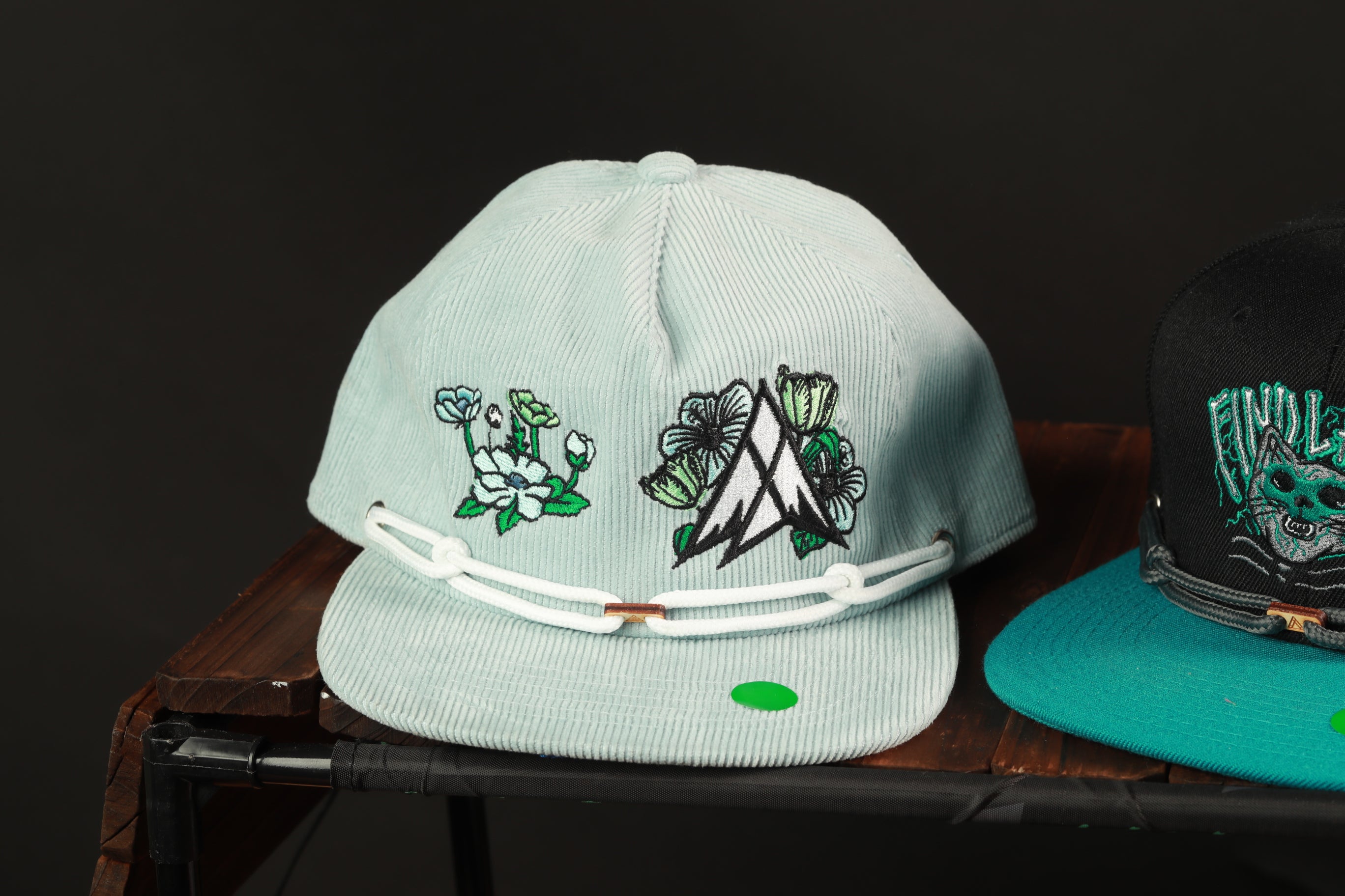two baseball caps on a wooden surface, one light blue and one teal, with floral and geometric designs on them.