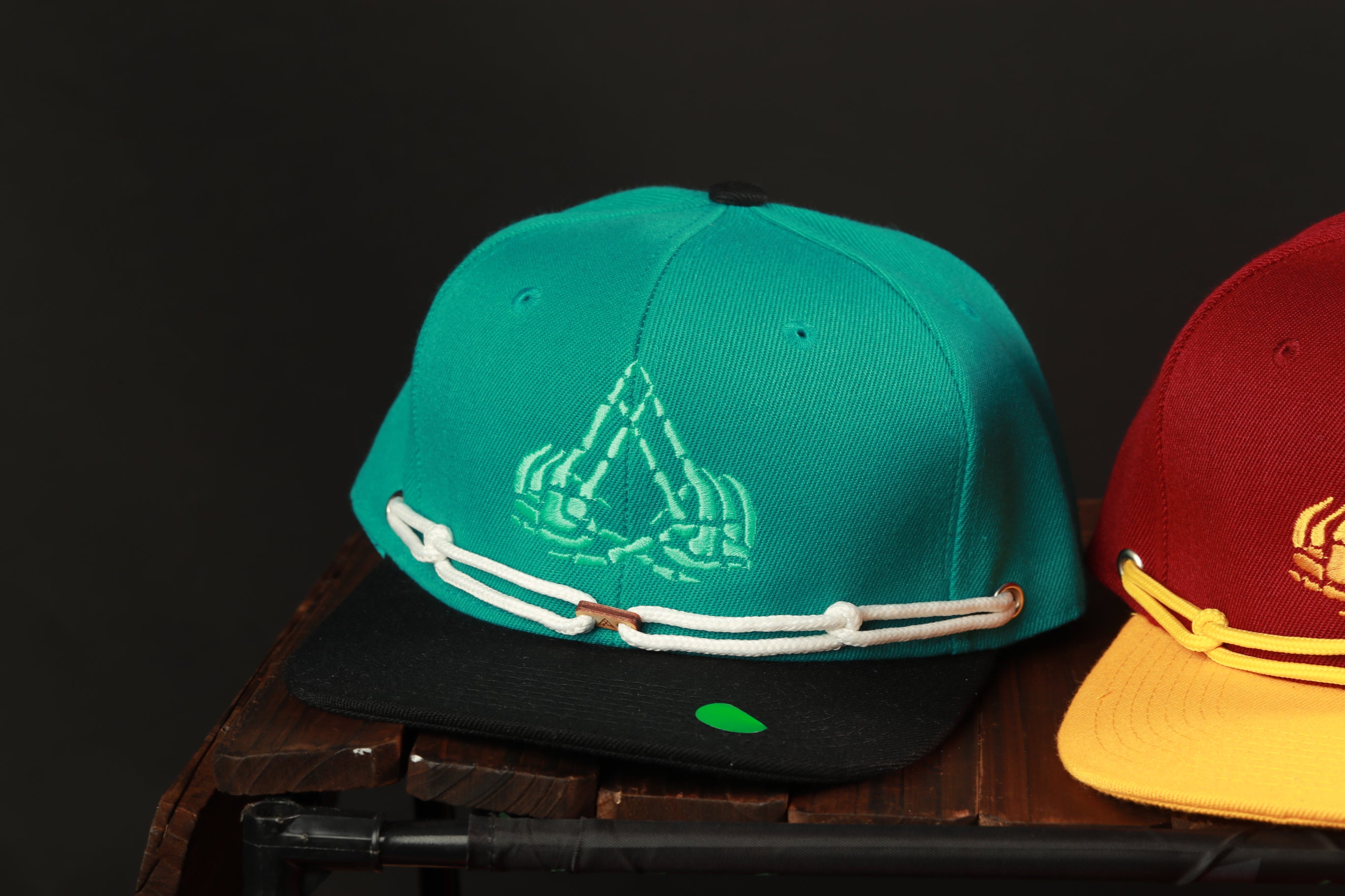 Two baseball caps are displayed on a wooden surface, one in a teal color and the other in a maroon color.