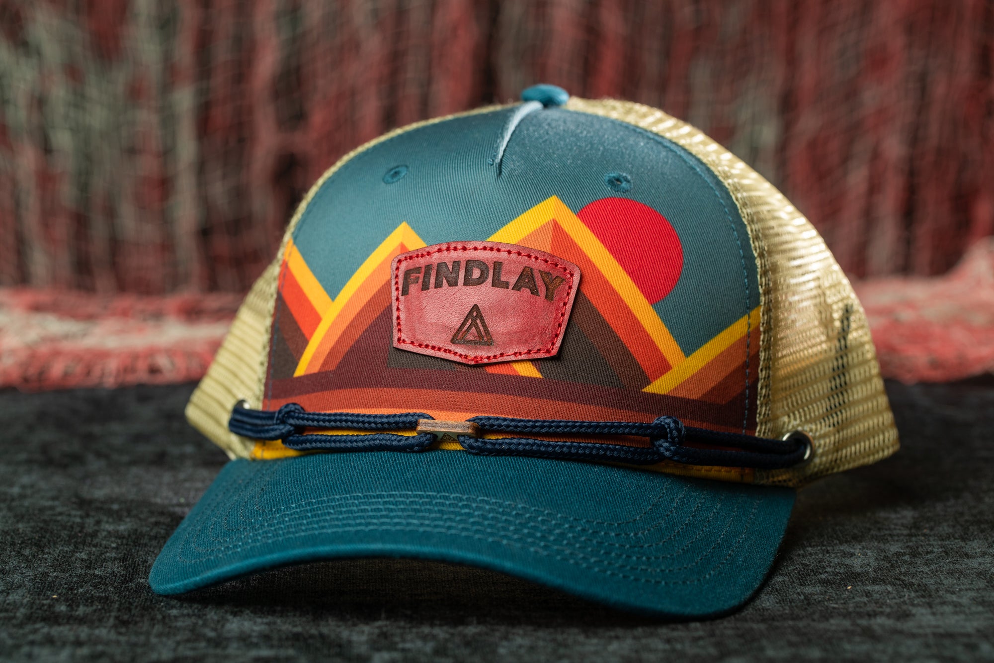 A colorful baseball cap with a mountain and sun design, featuring the word "FINDLAY" on the front.