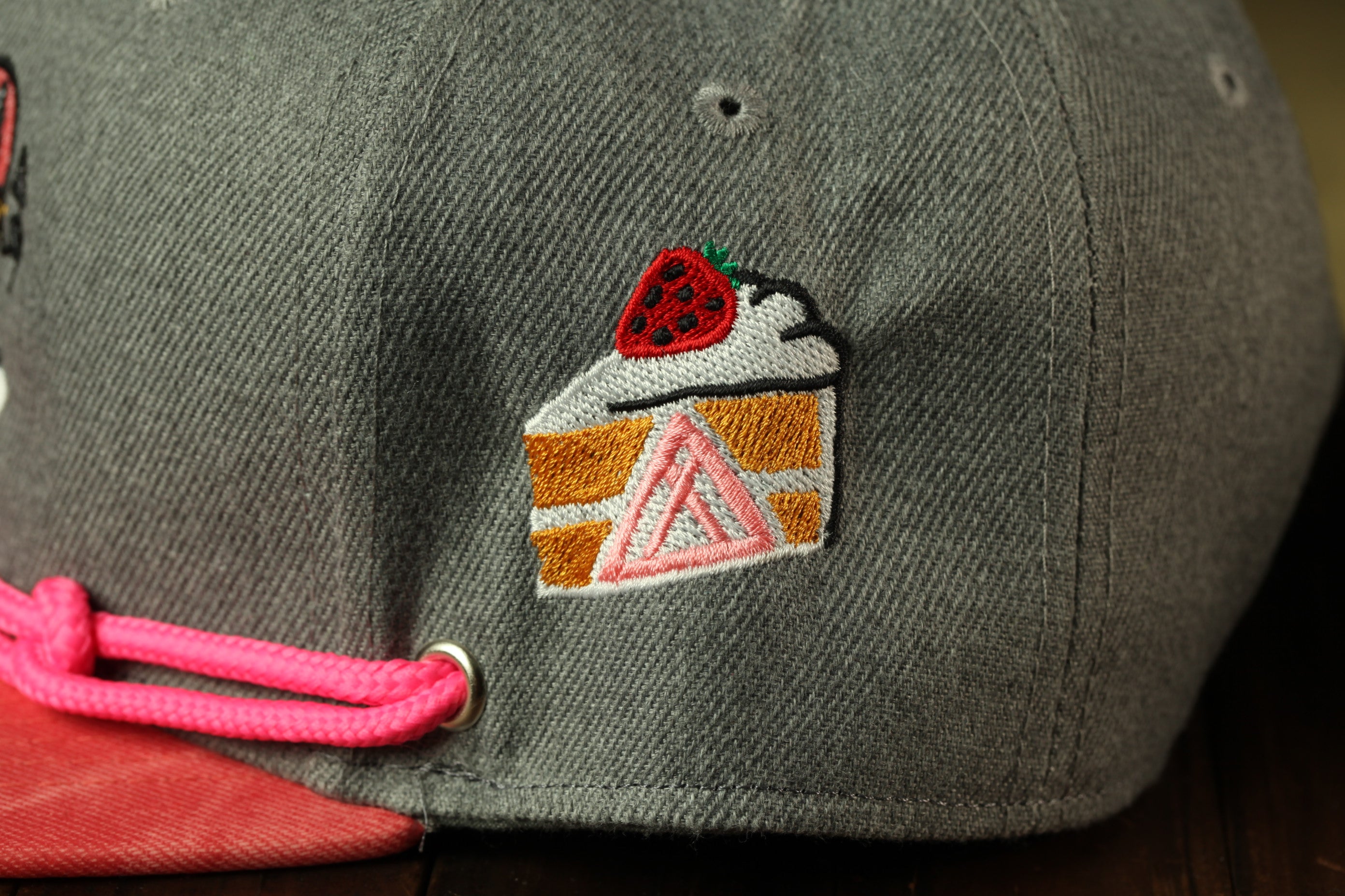 A close-up view of a gray baseball cap with a pink string and a small embroidered design depicting a slice of cake with a strawberry on top.