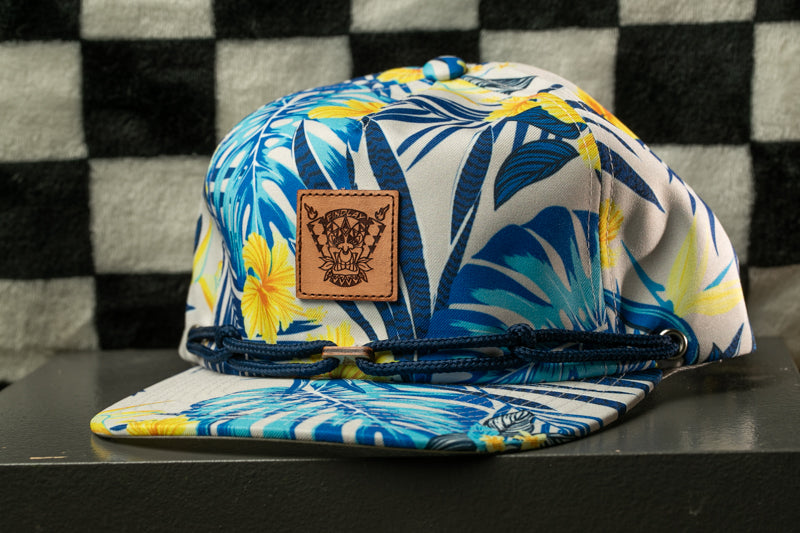 A colorful tropical-themed baseball cap with a leather patch on the front, displayed on a black surface against a checkered background.