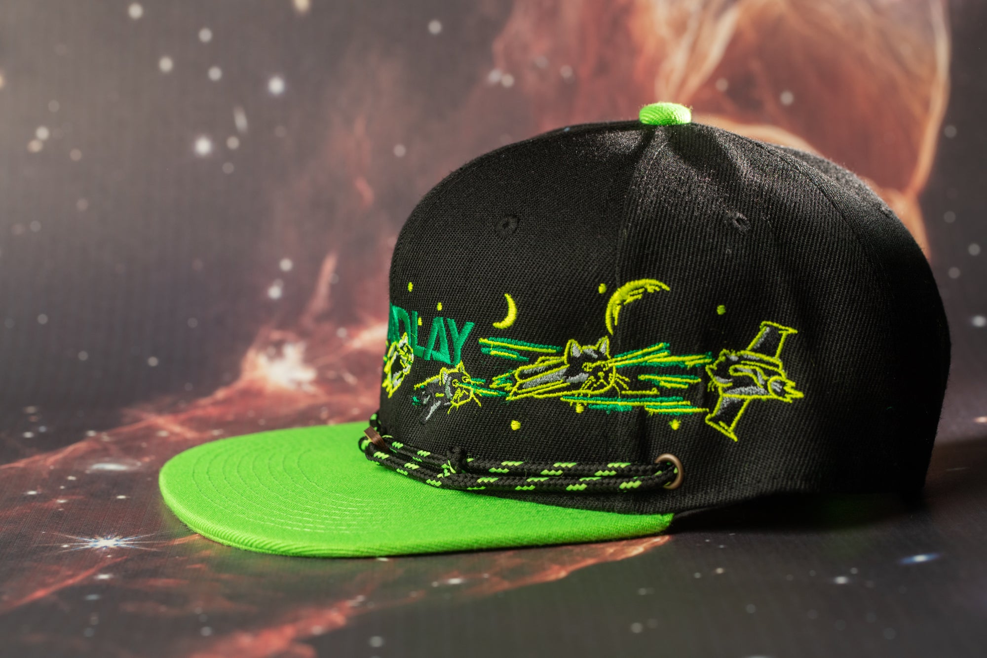 A black baseball cap with a green brim and a design featuring a space-themed illustration, including a rocket ship and a crescent moon.