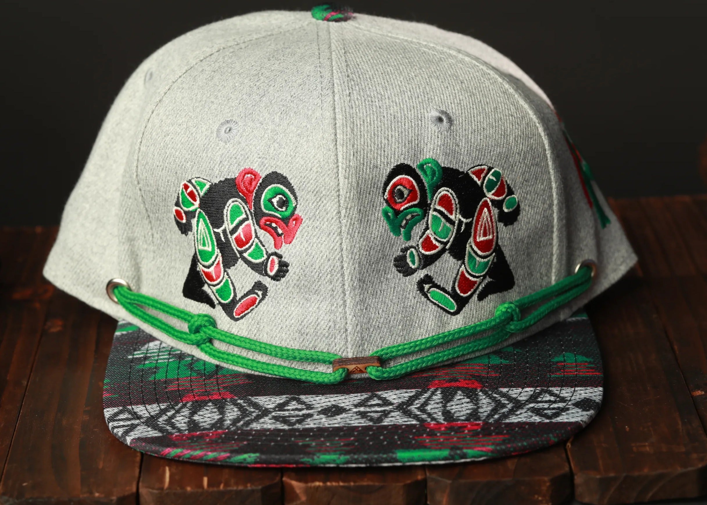 A gray baseball cap with colorful designs on the front, including two stylized animal faces, is displayed on a wooden surface.