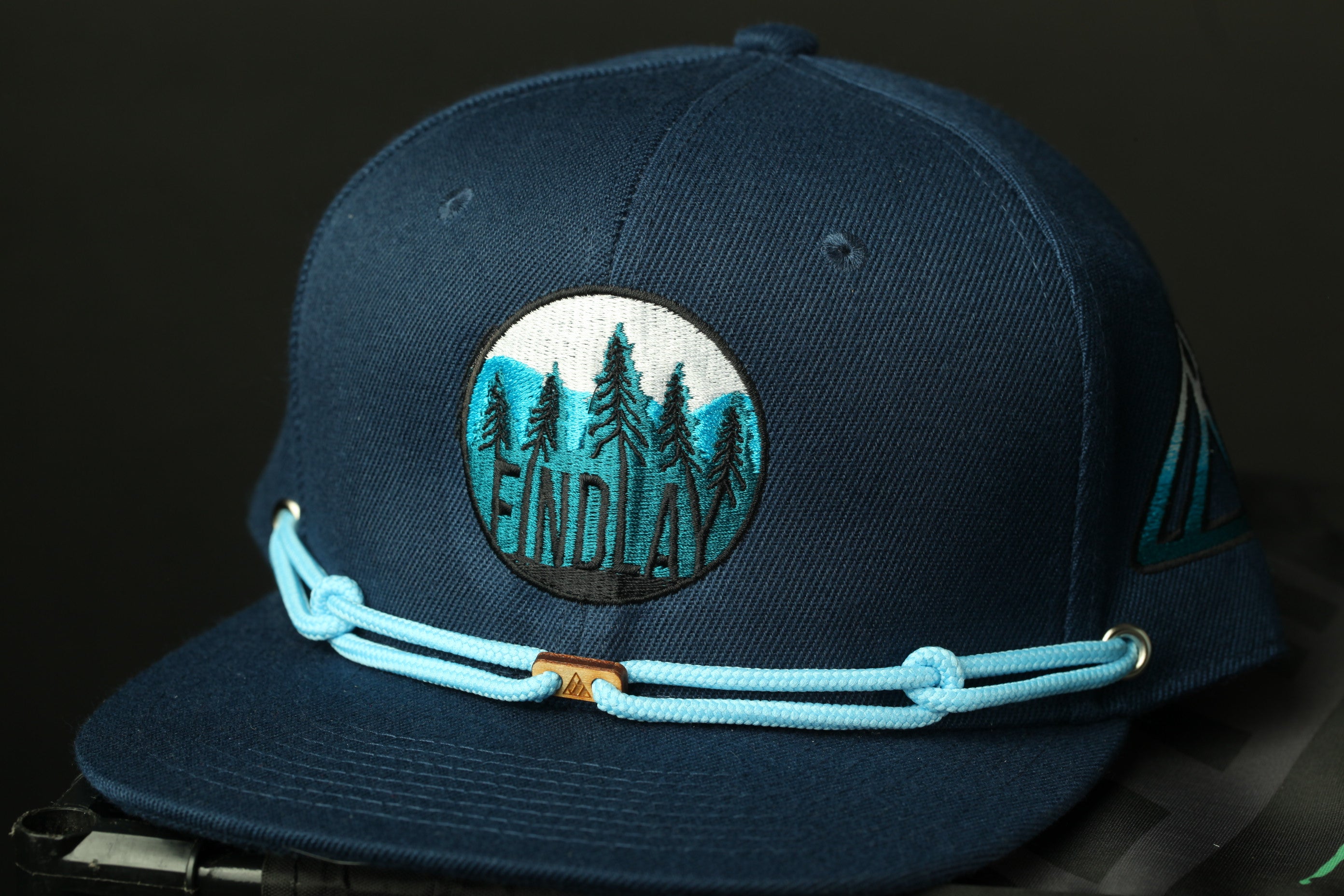 A navy blue baseball cap with a circular embroidered logo on the front, featuring a mountain landscape with pine trees.