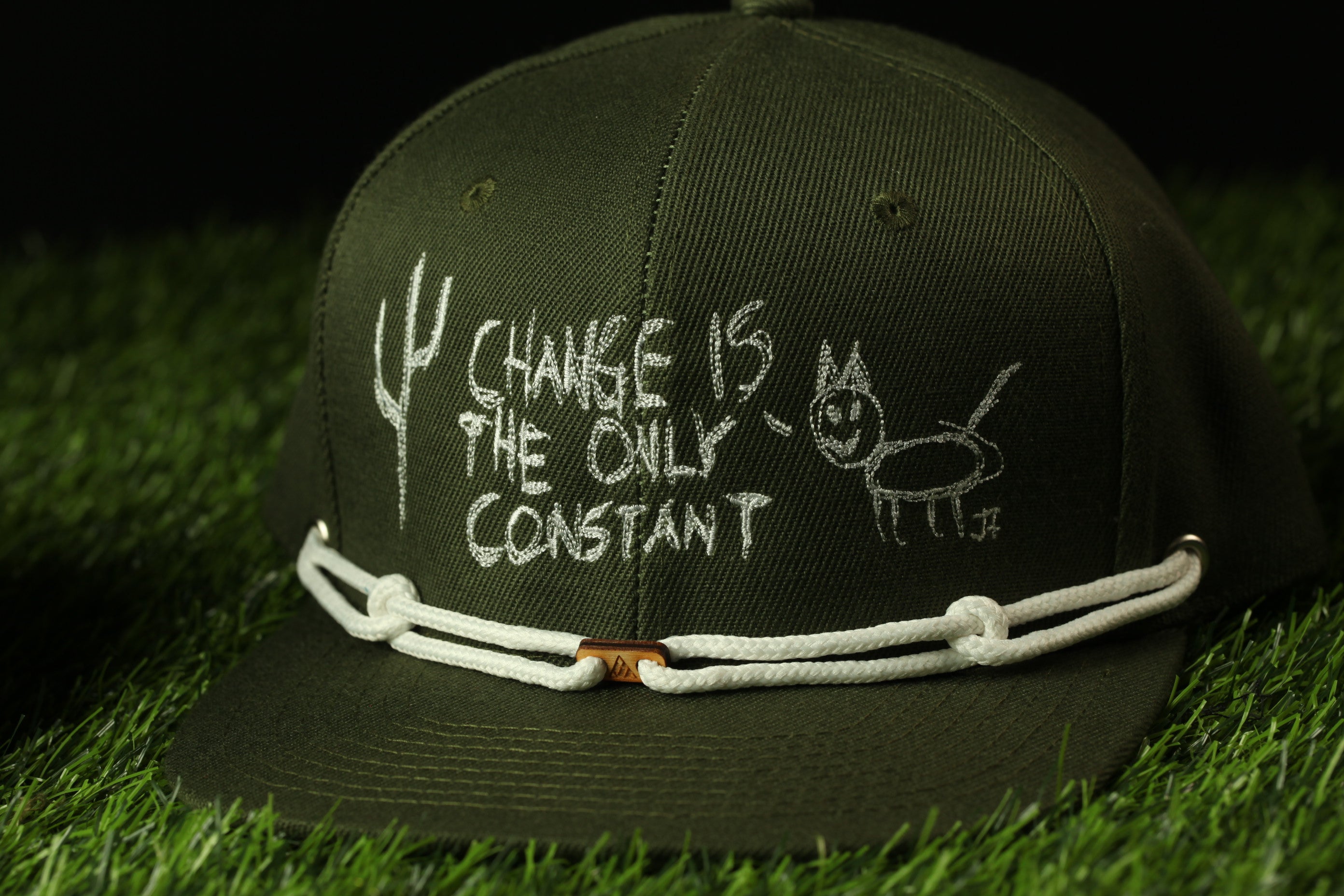 A green baseball cap with white text and drawings on it, placed on a grassy surface.