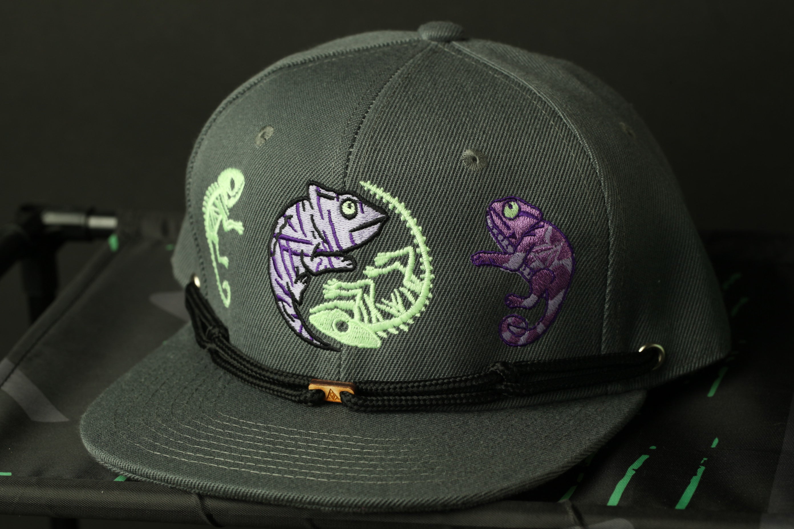 A gray baseball cap with a unique design featuring two chameleons in purple and green colors.