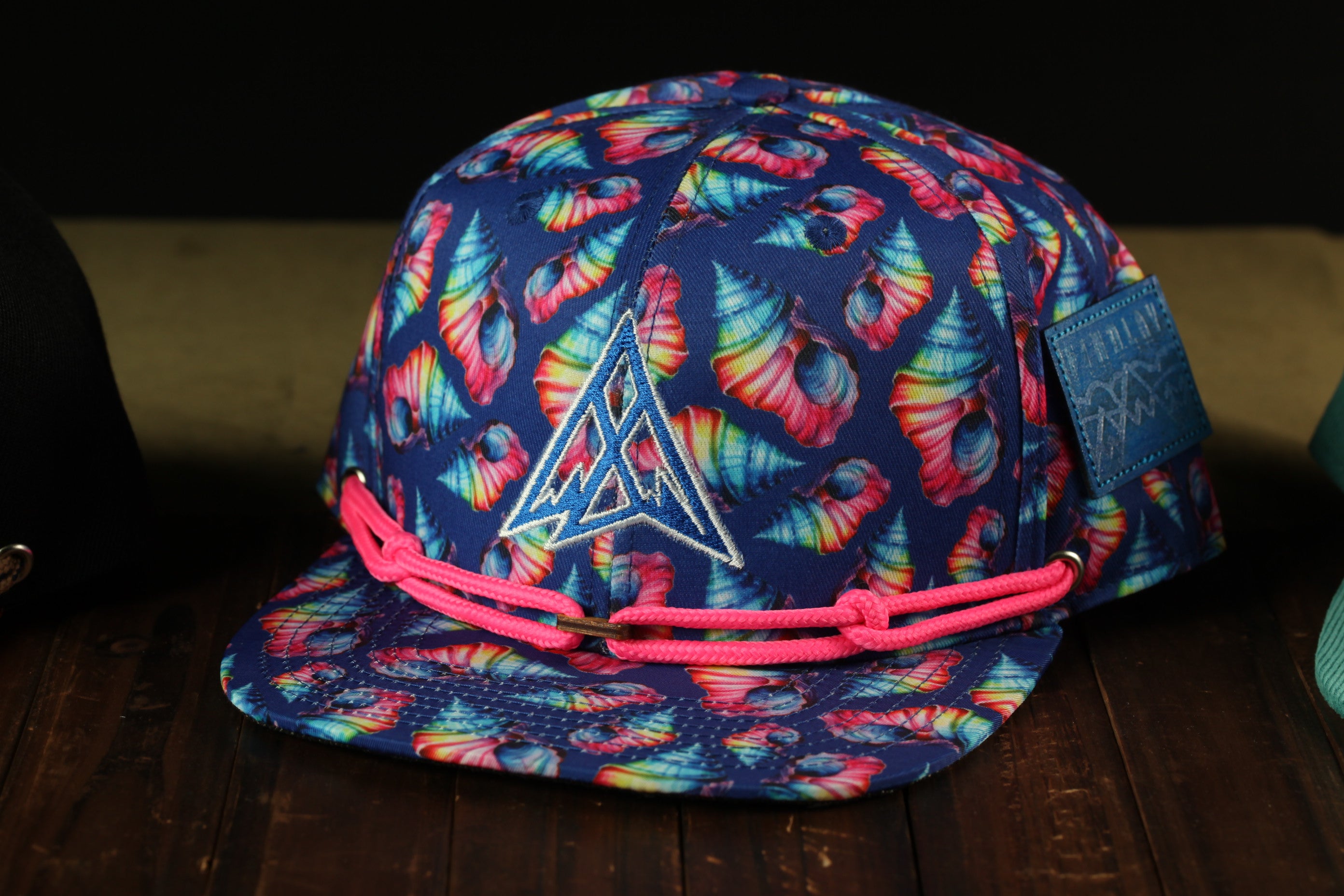 A colorful, tie-dye baseball cap with a pattern of seashells and a triangular logo on the front, sitting on a wooden surface.