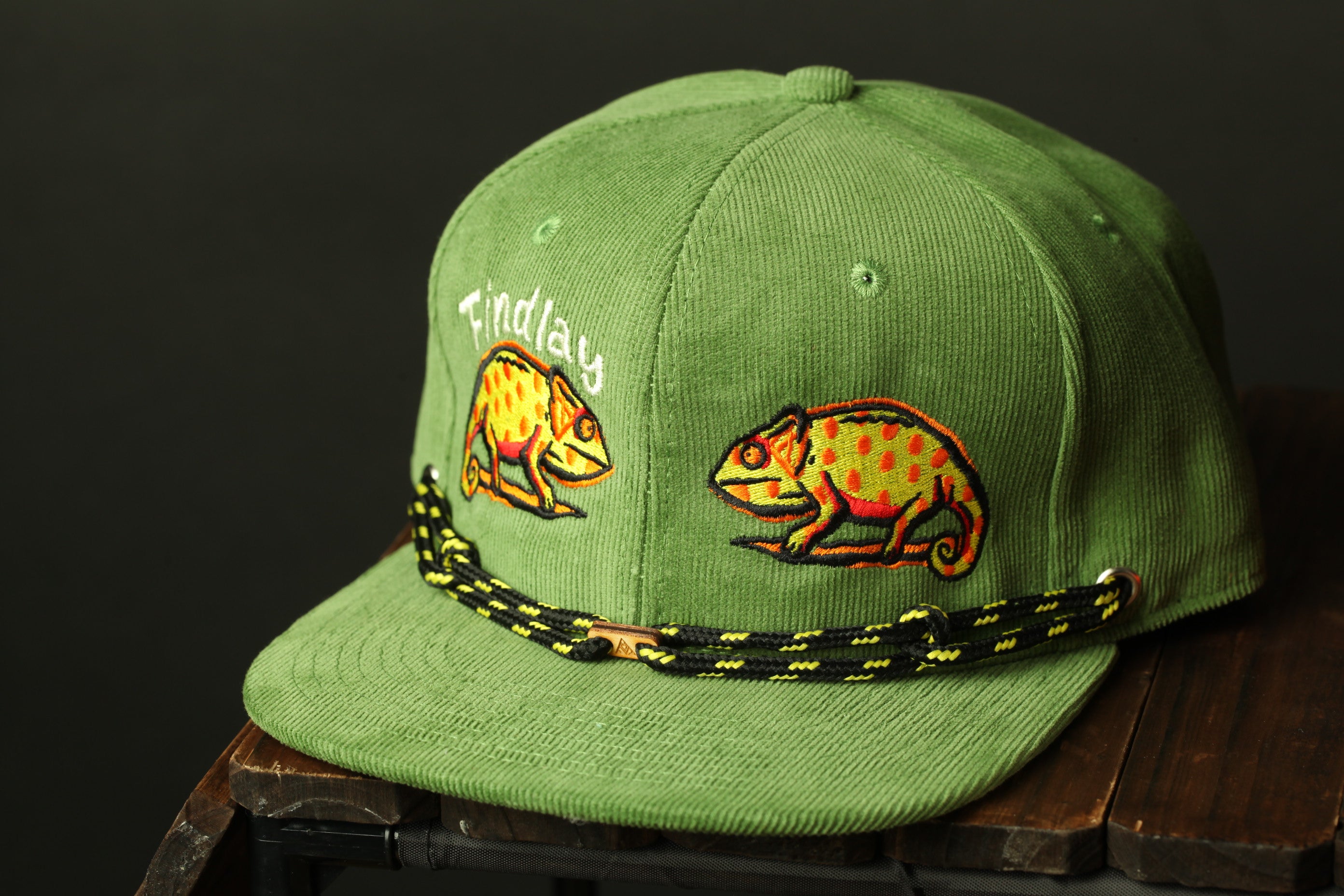 A green baseball cap with a black and yellow braided strap, featuring two chameleon illustrations on the front.