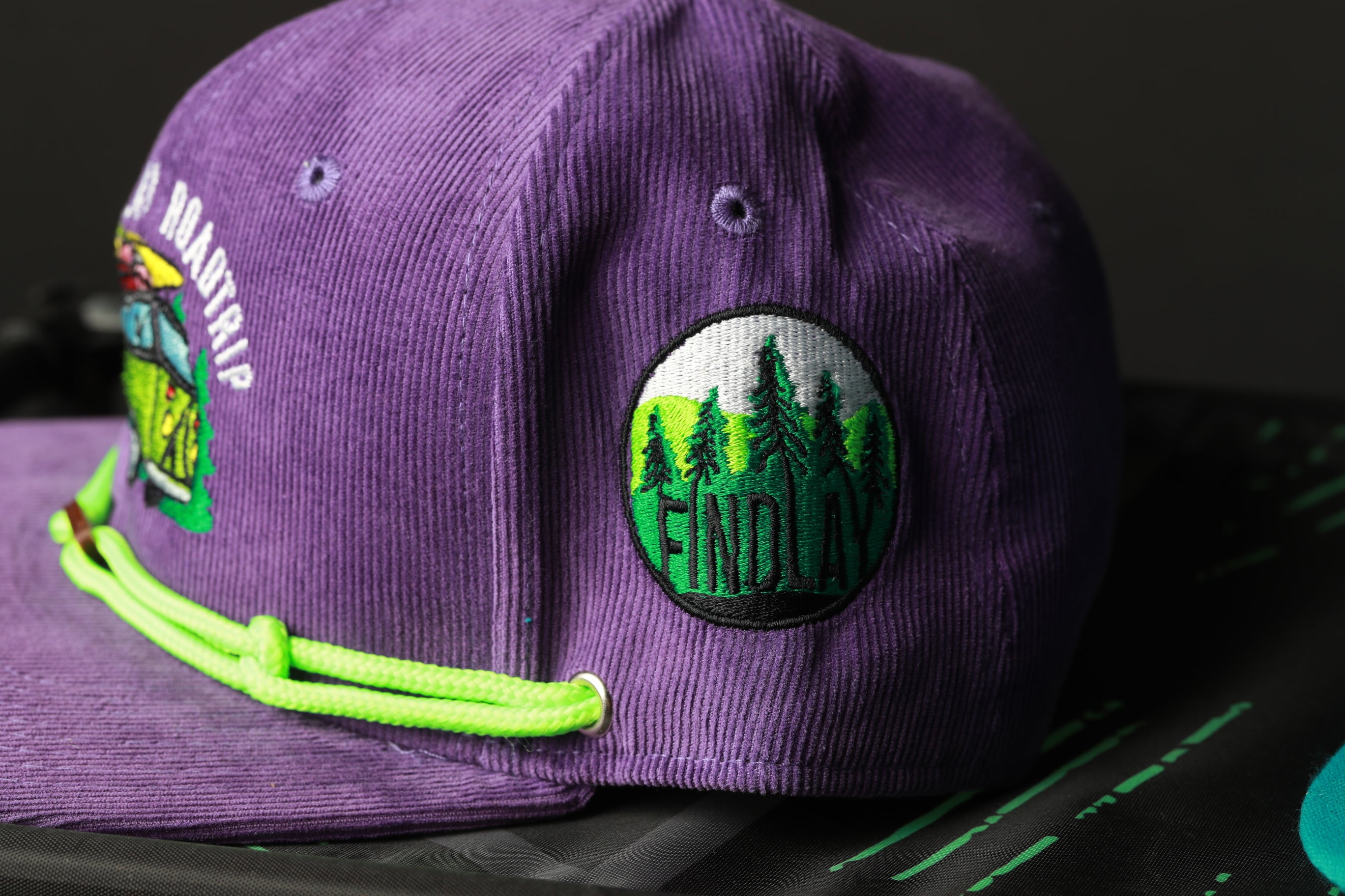 A purple baseball cap with a green string and a patch on the front that says "FINDlay".