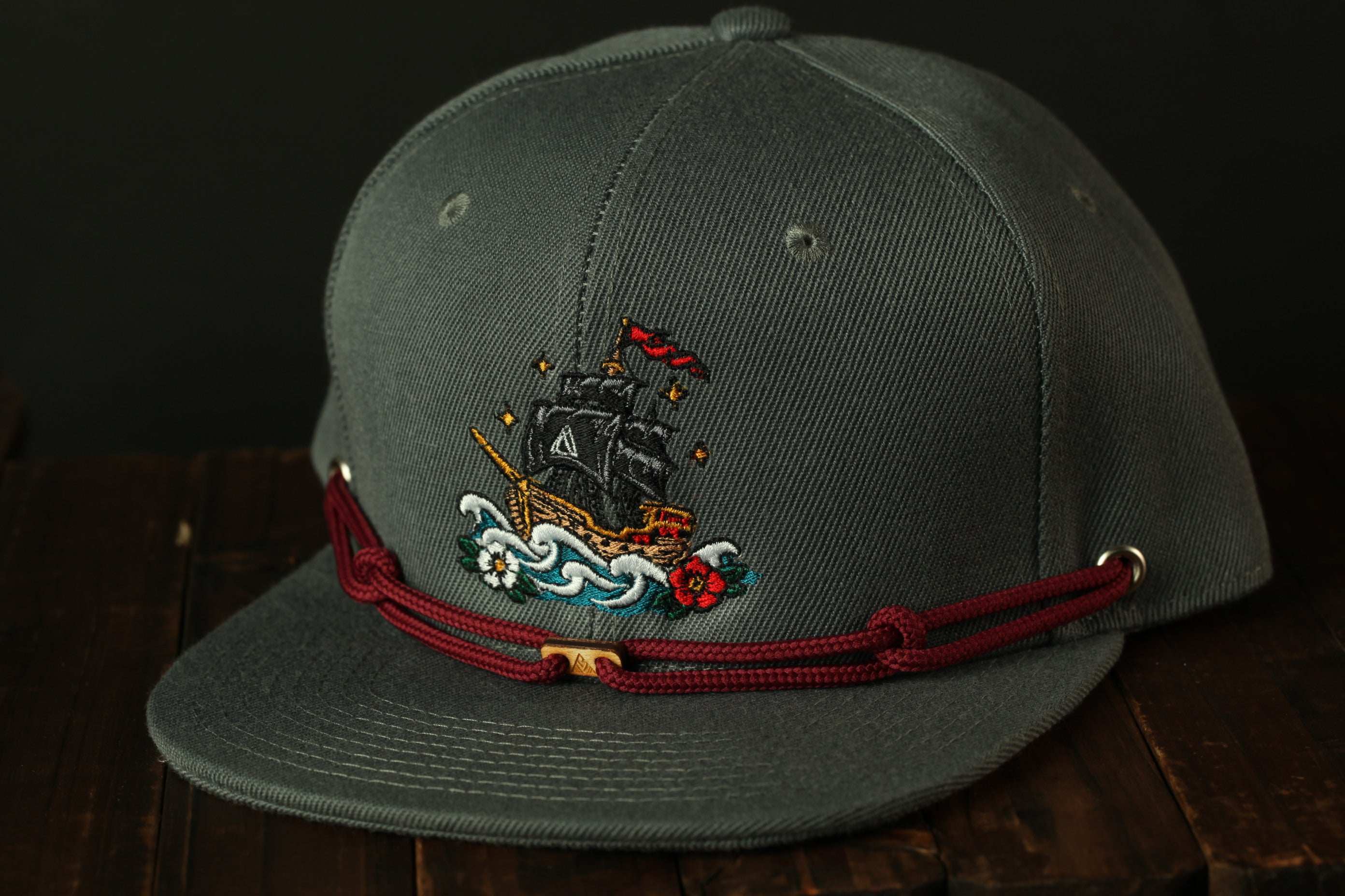 A gray baseball cap with a red string and a ship embroidered on the front, resting on a wooden surface.
