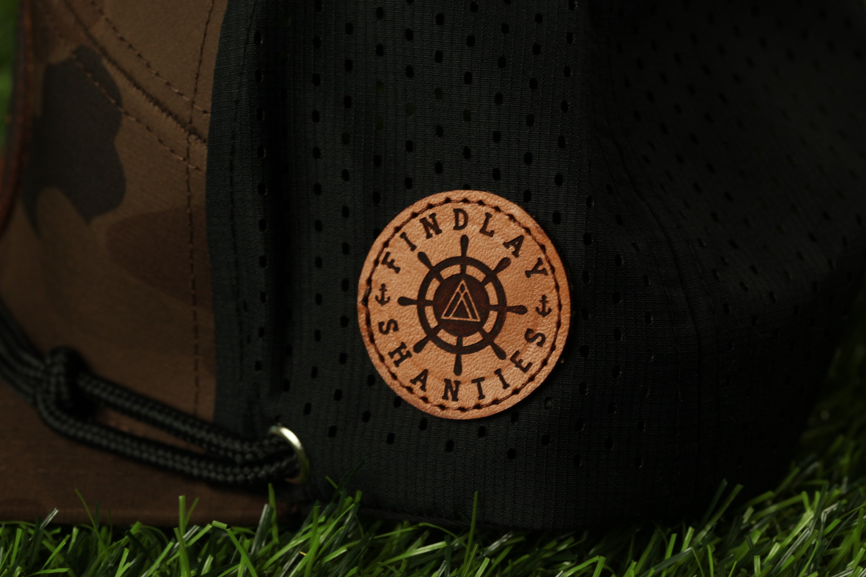 A close-up view of a black and brown baseball cap with a circular leather patch on the front, featuring a ship's wheel design and the text "FIND LAYS" printed on it.