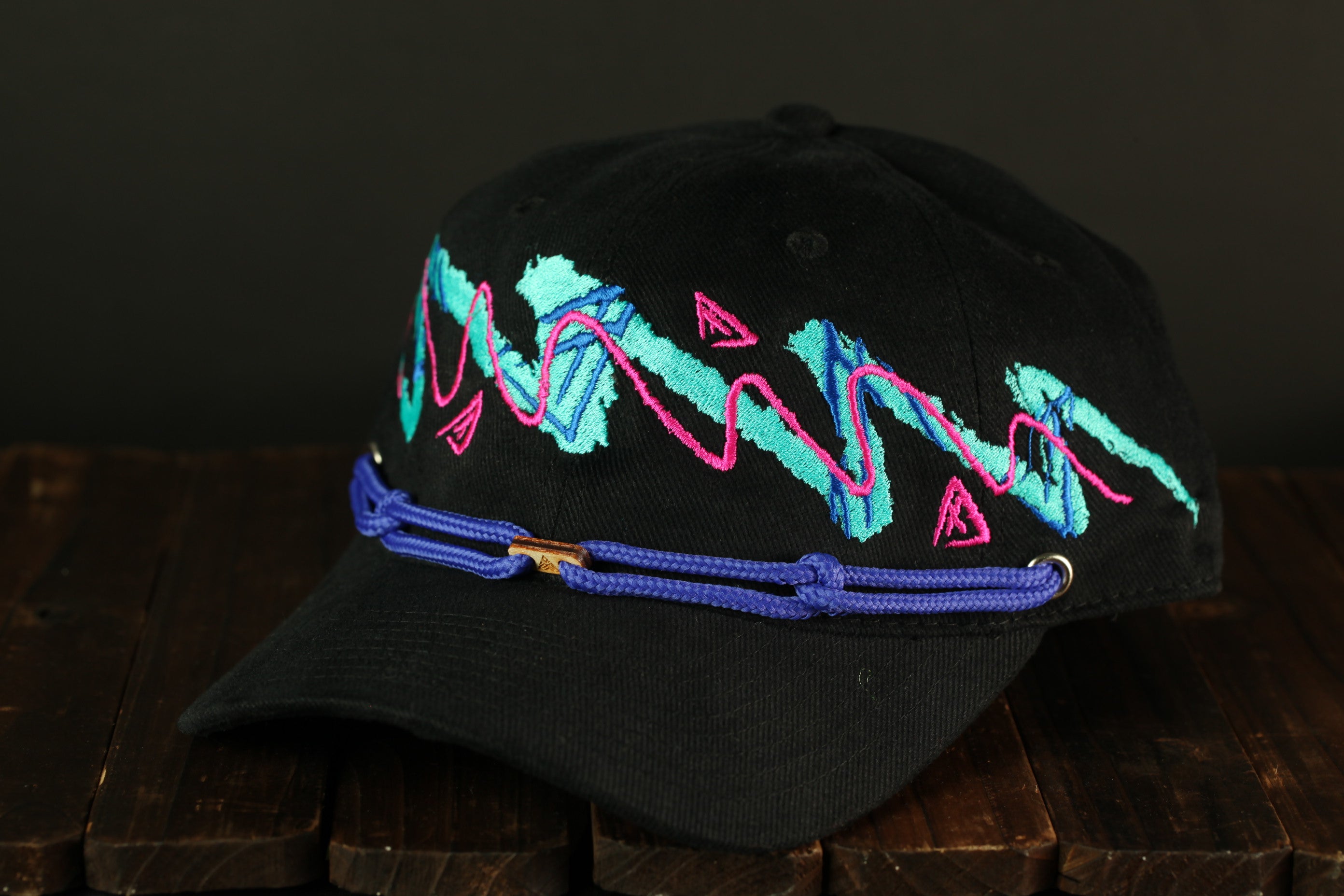 A black baseball cap with a colorful, abstract embroidered design on the front, featuring a mix of pink, blue, and green lines and shapes.