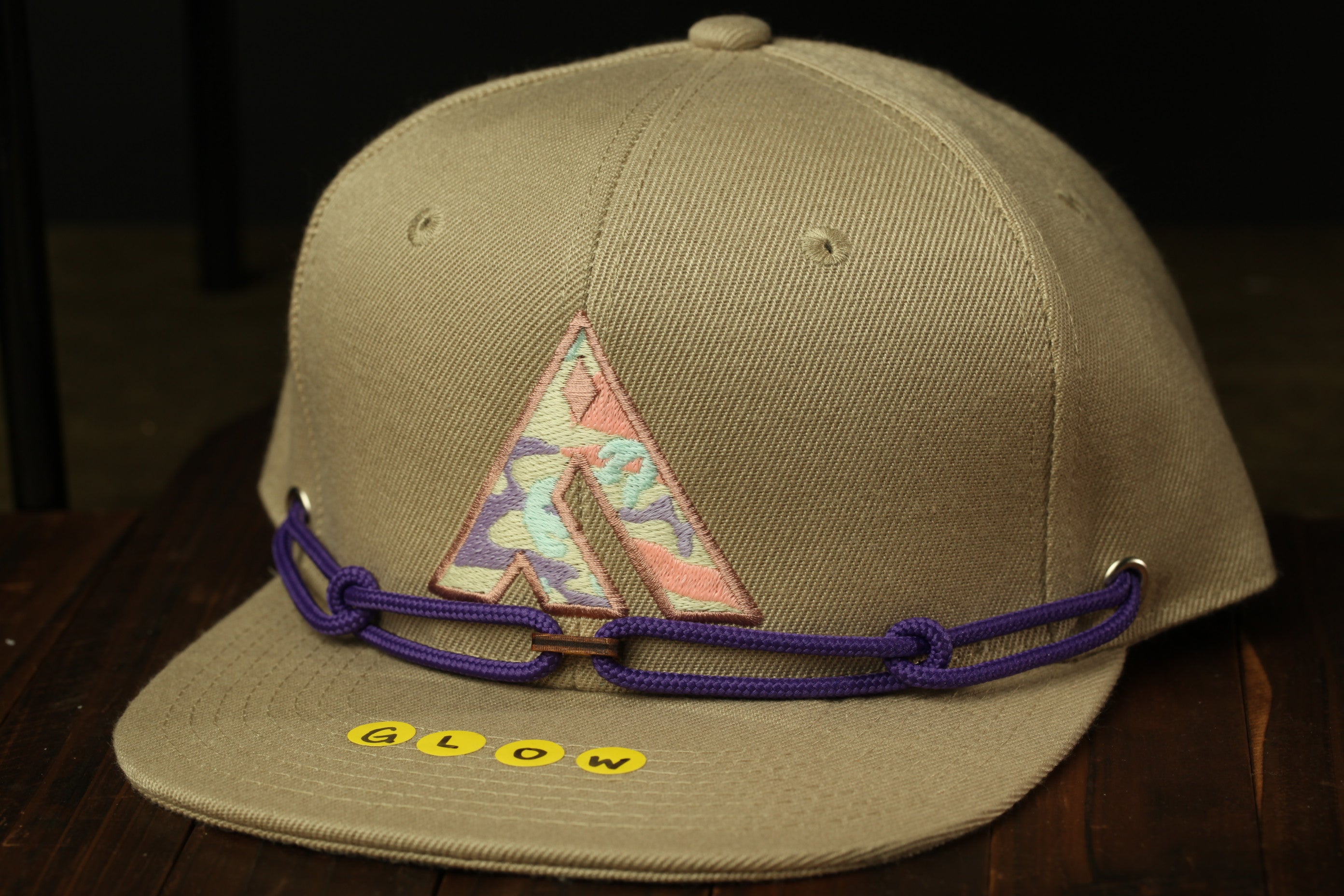 A tan baseball cap with a pink and blue floral pattern and a purple rope strap, sitting on a wooden surface.