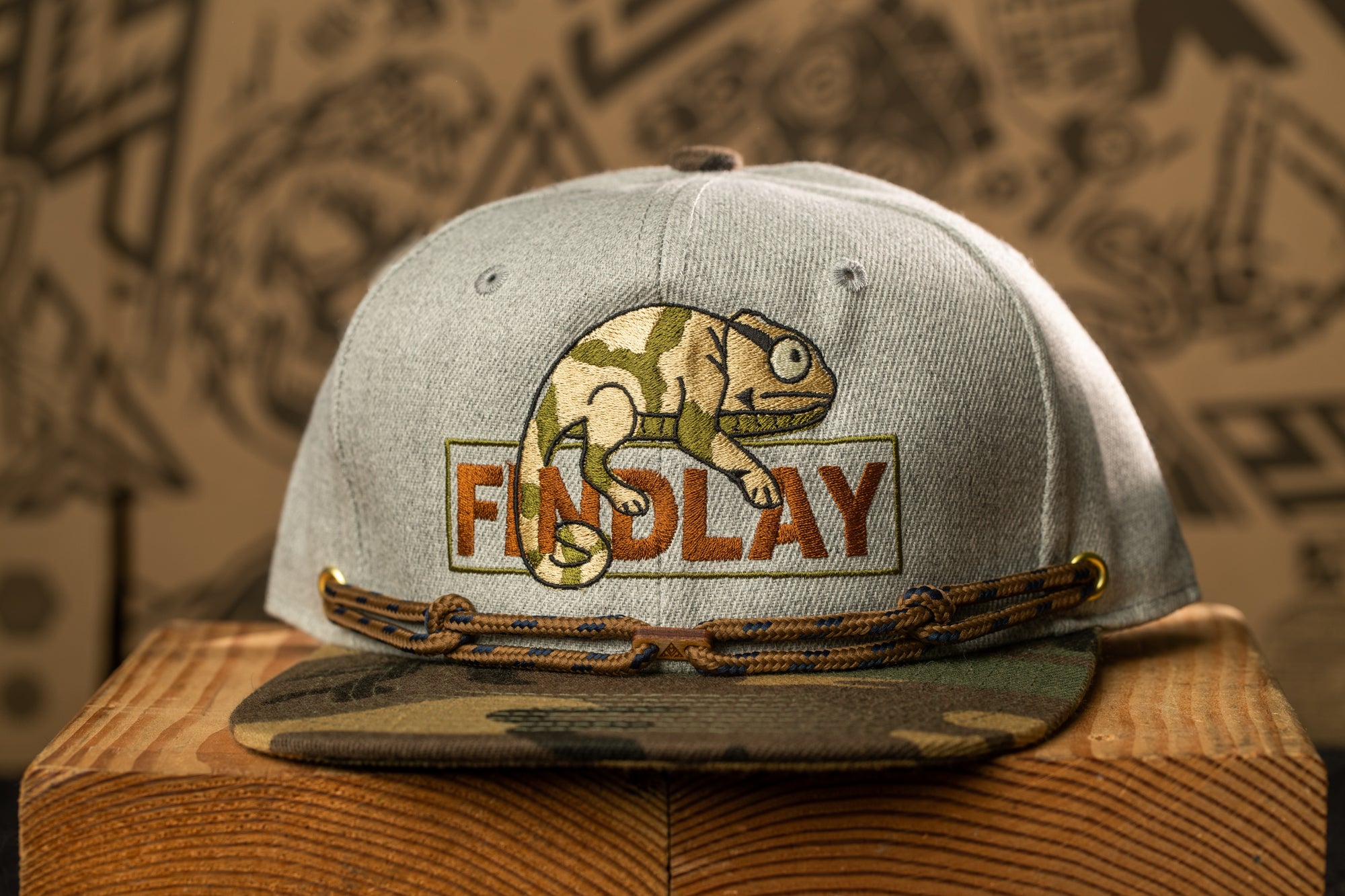 A gray baseball cap with a camouflage pattern and a cartoon chameleon on it, sitting on a wooden surface.