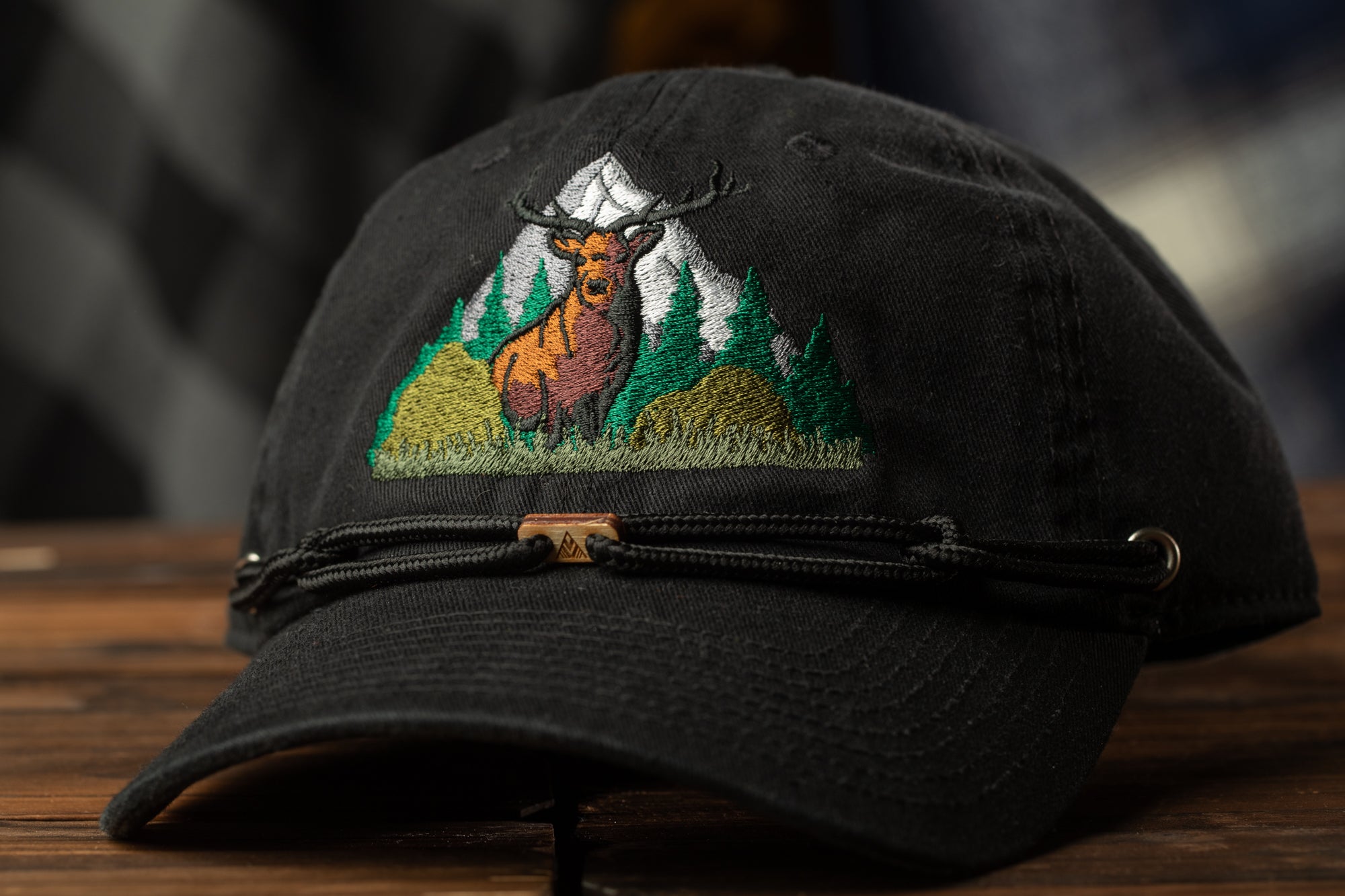 A black baseball cap with a detailed embroidered design of a deer standing in a forest landscape.