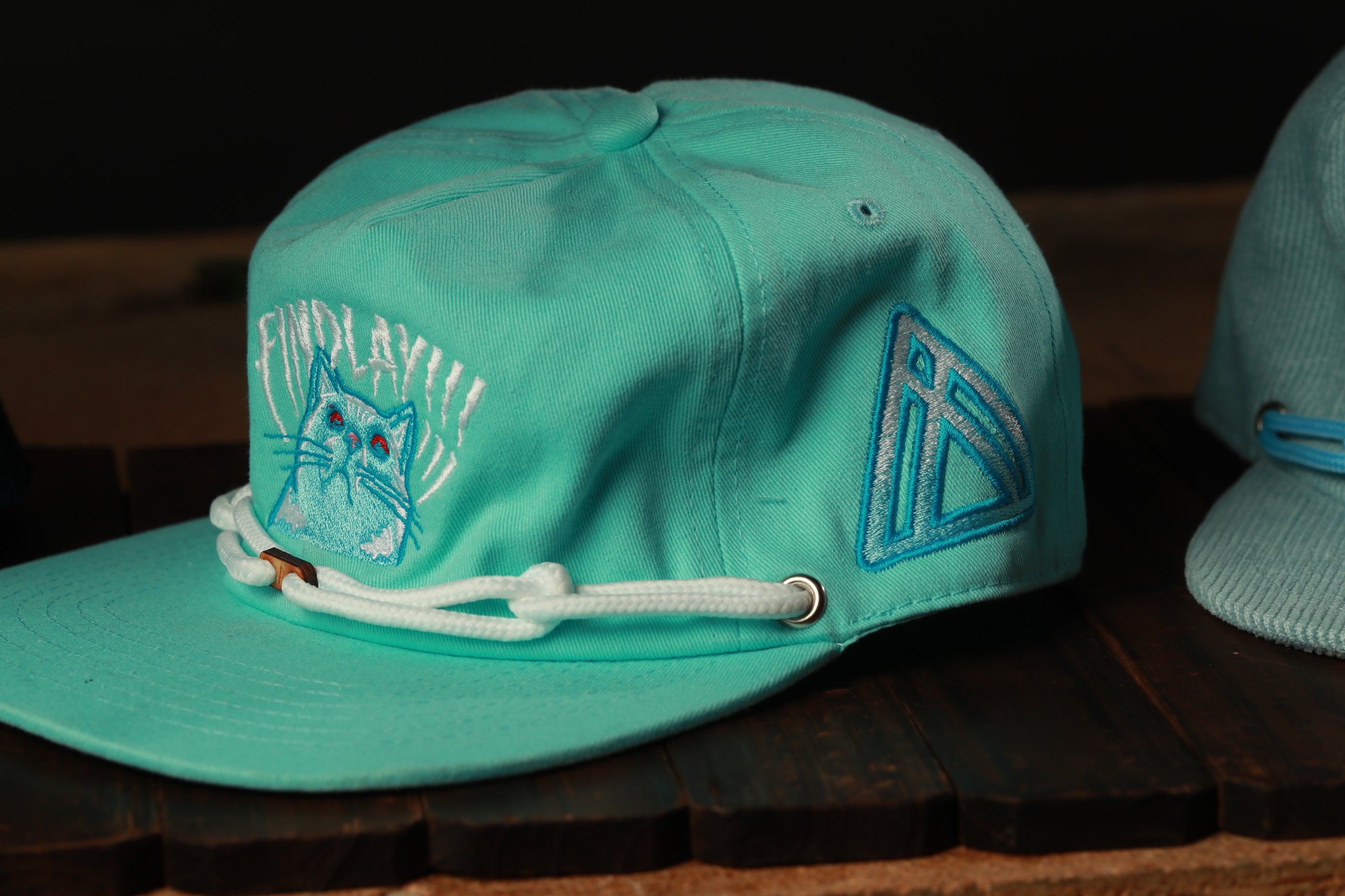 A turquoise baseball cap with a cat embroidered on it, featuring a white lace strap and a blue triangle logo on the side.