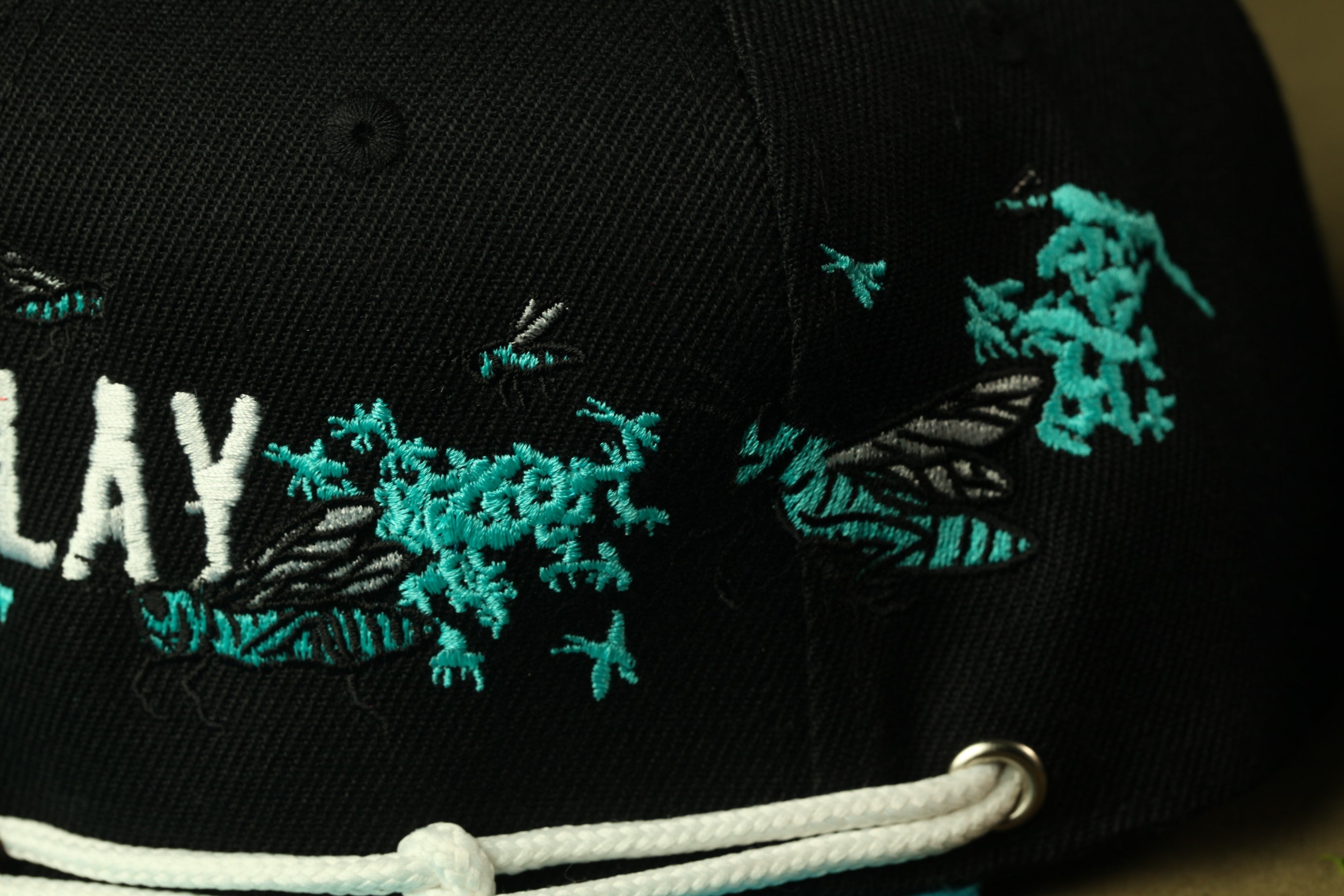 a close-up view of a black baseball cap with a detailed embroidered design featuring a bee and other insects.