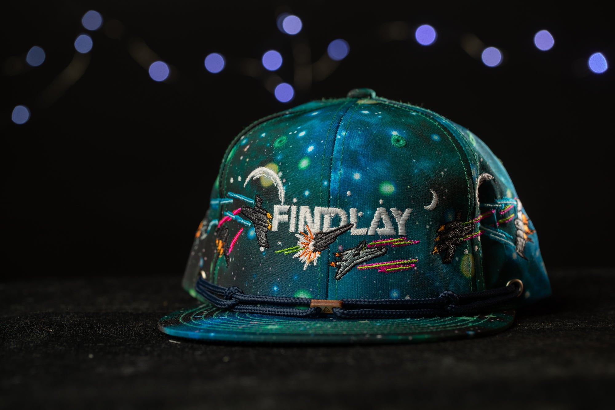 A colorful, space-themed baseball cap with the word "FINDlay" printed on it, set against a dark background with twinkling lights.