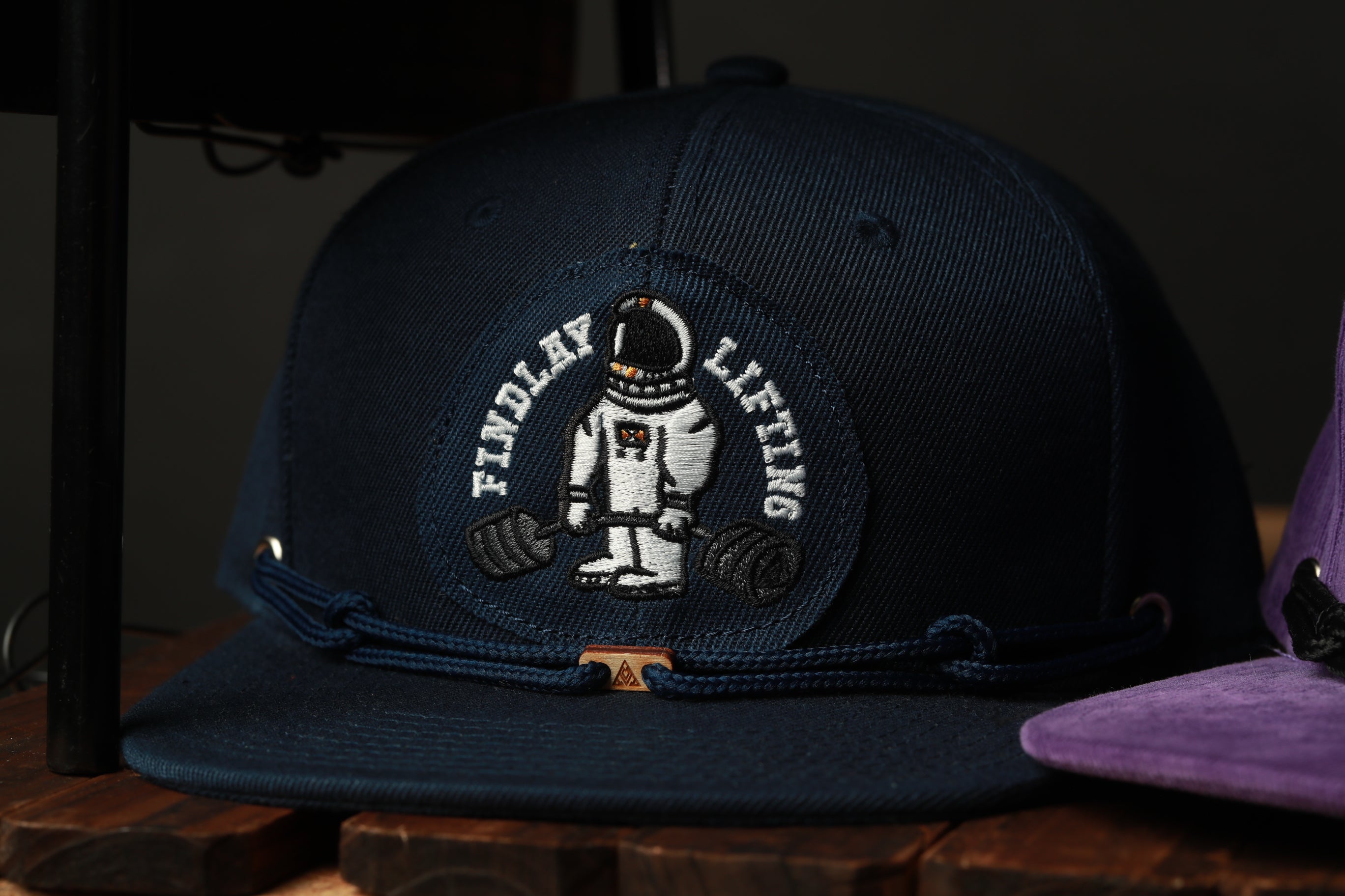 A navy blue baseball cap with a white and black embroidered design featuring a space suit and the text "FIND A LIFTING" on the front.