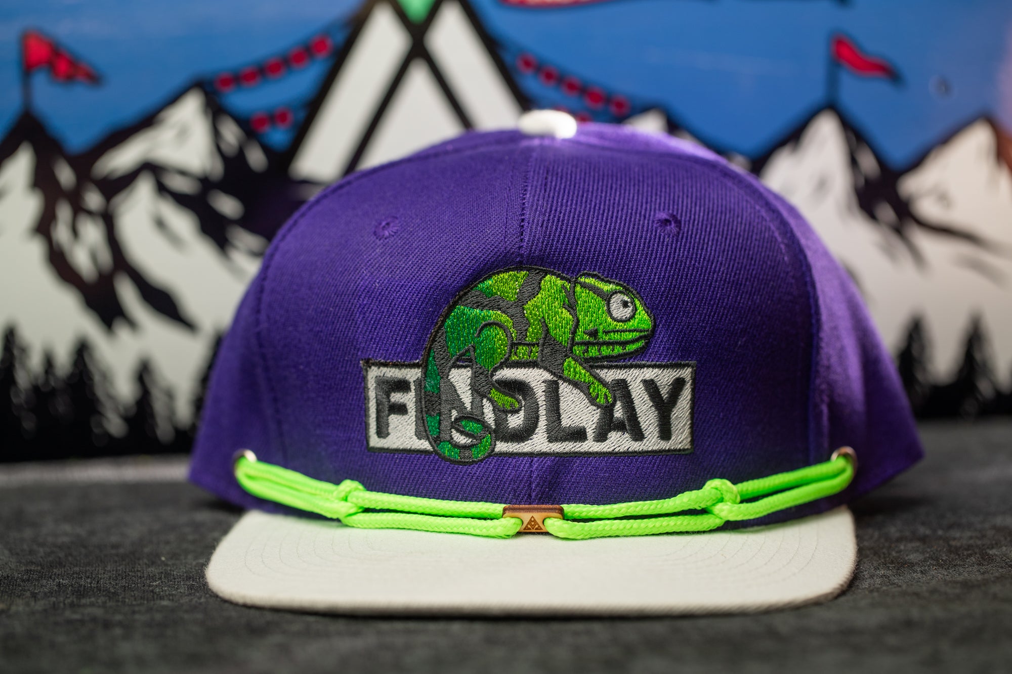 A purple baseball cap with a green chameleon design and the word "FADLAY" printed on it, along with a green string or strap.