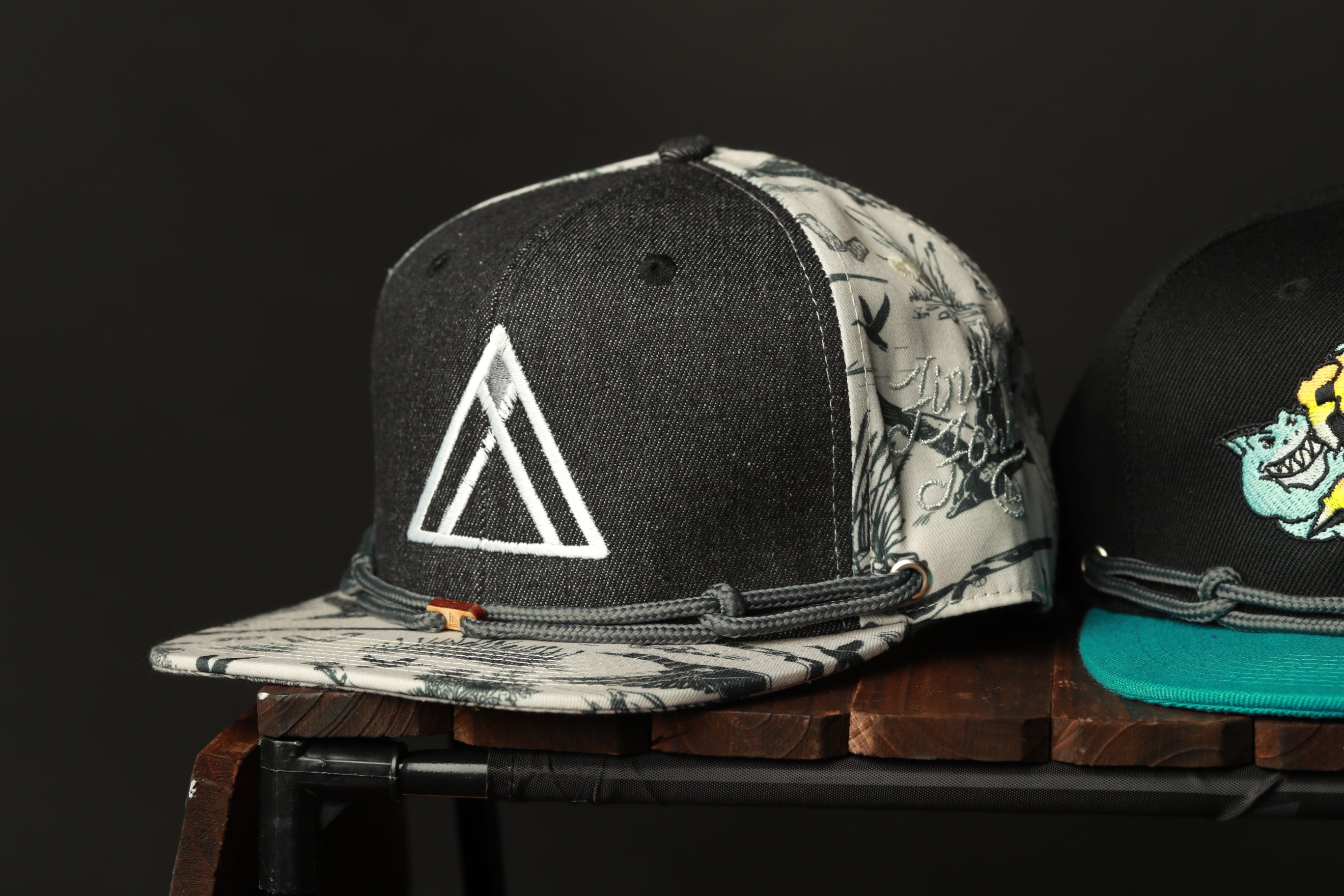 Two baseball caps are displayed on a wooden surface, one with a black and white pattern and the other with a teal and black pattern.