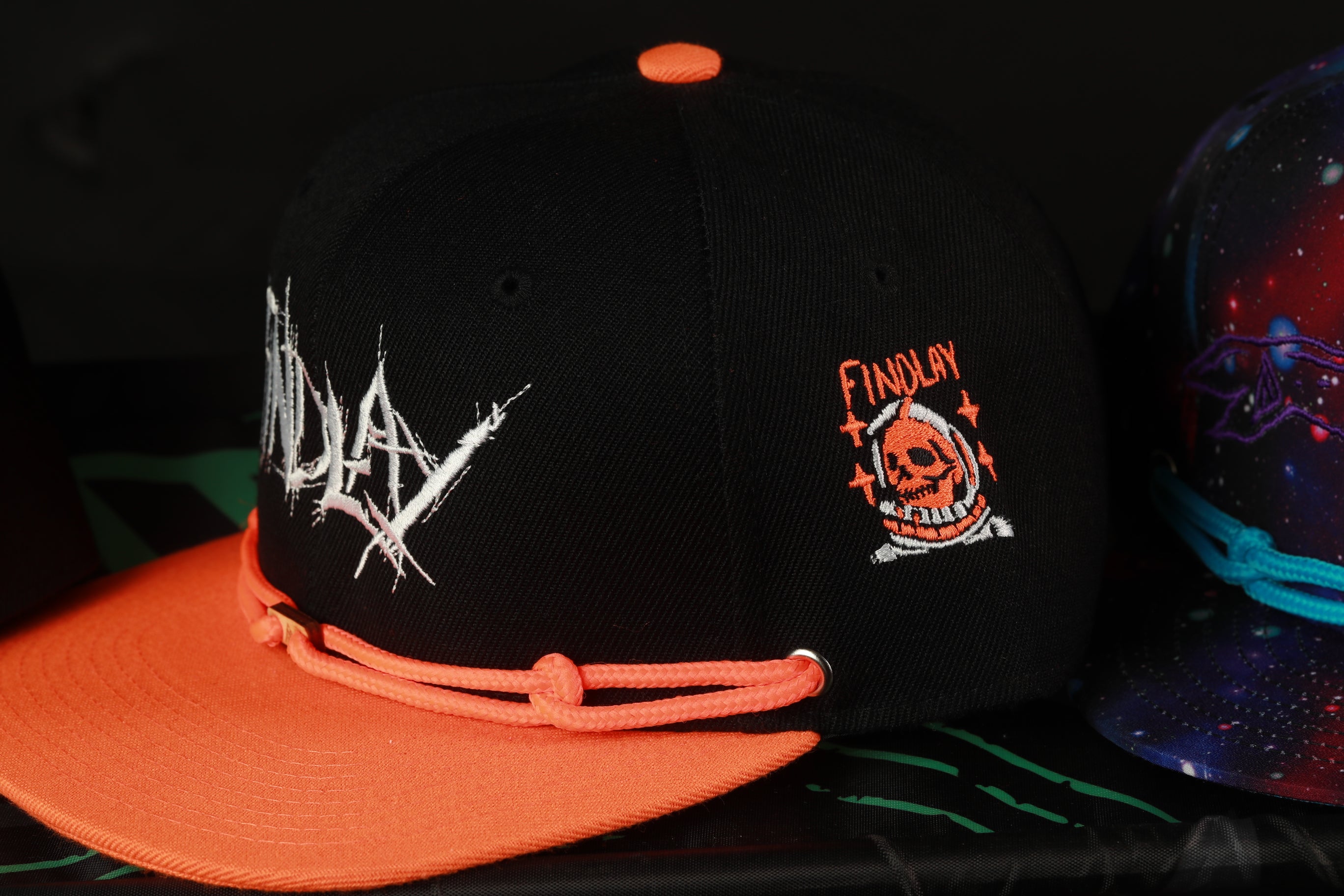 A black baseball cap with an orange brim and a skull logo on the side, along with a matching orange strap.