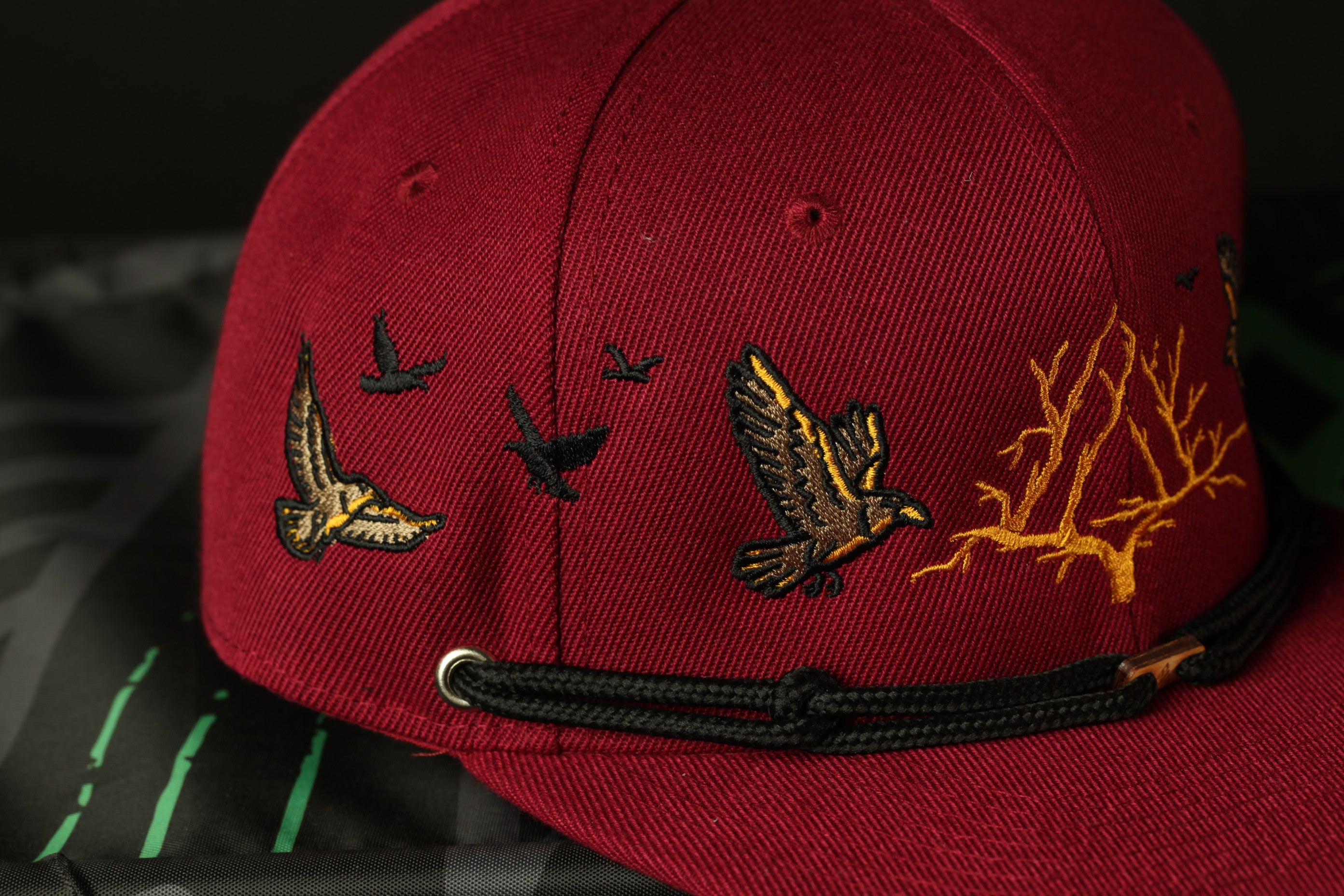 A close-up view of a maroon baseball cap with a black strap and embroidered designs of birds in flight.