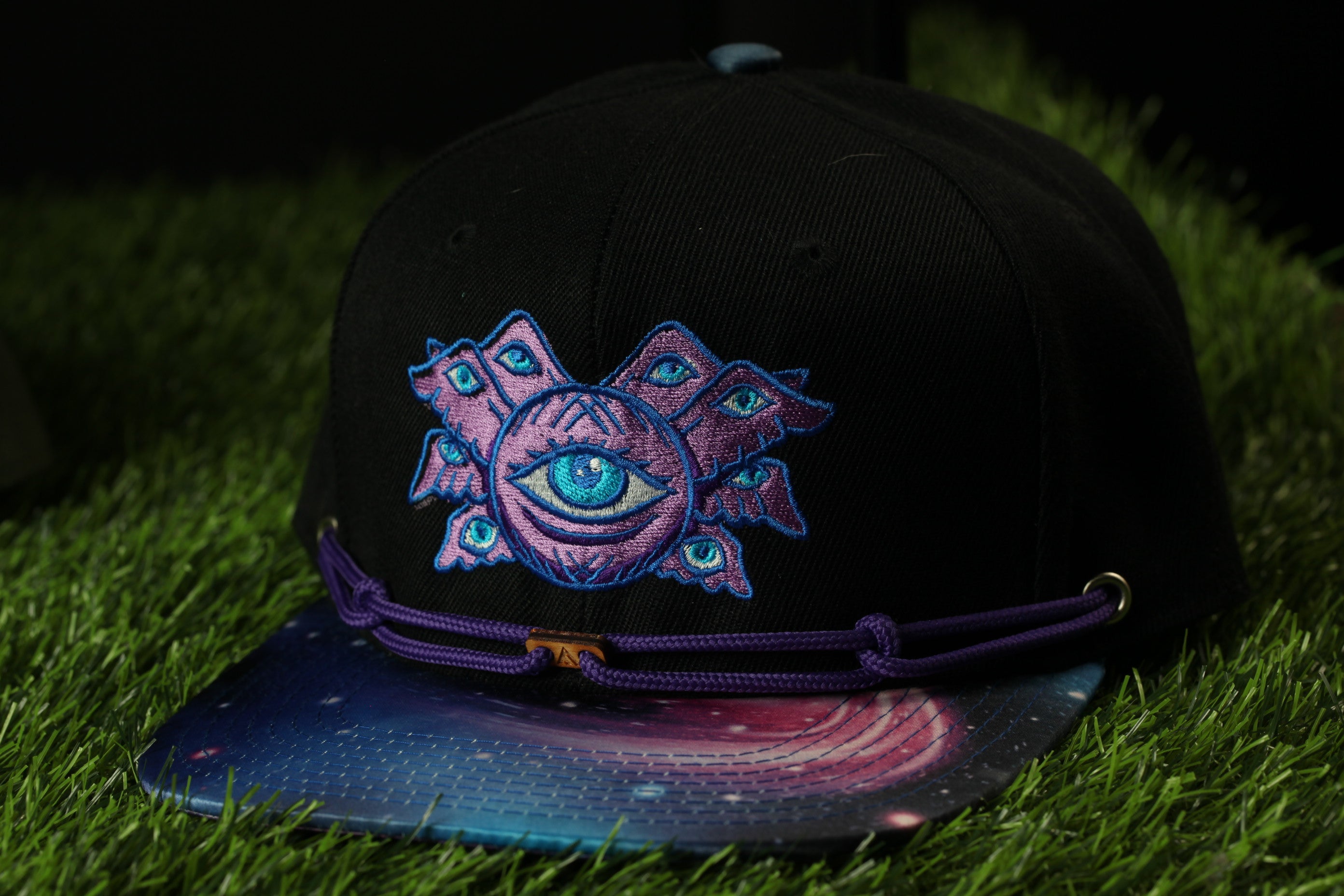 A black baseball cap with a colorful, abstract design on the front, featuring a large eye-like symbol surrounded by smaller geometric shapes in shades of purple and blue.